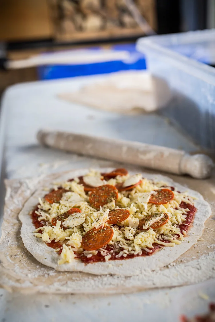 Unbaked pizza with pepperoni, cheese, and herbs on a lightly floured surface with a rolling pin in the background.