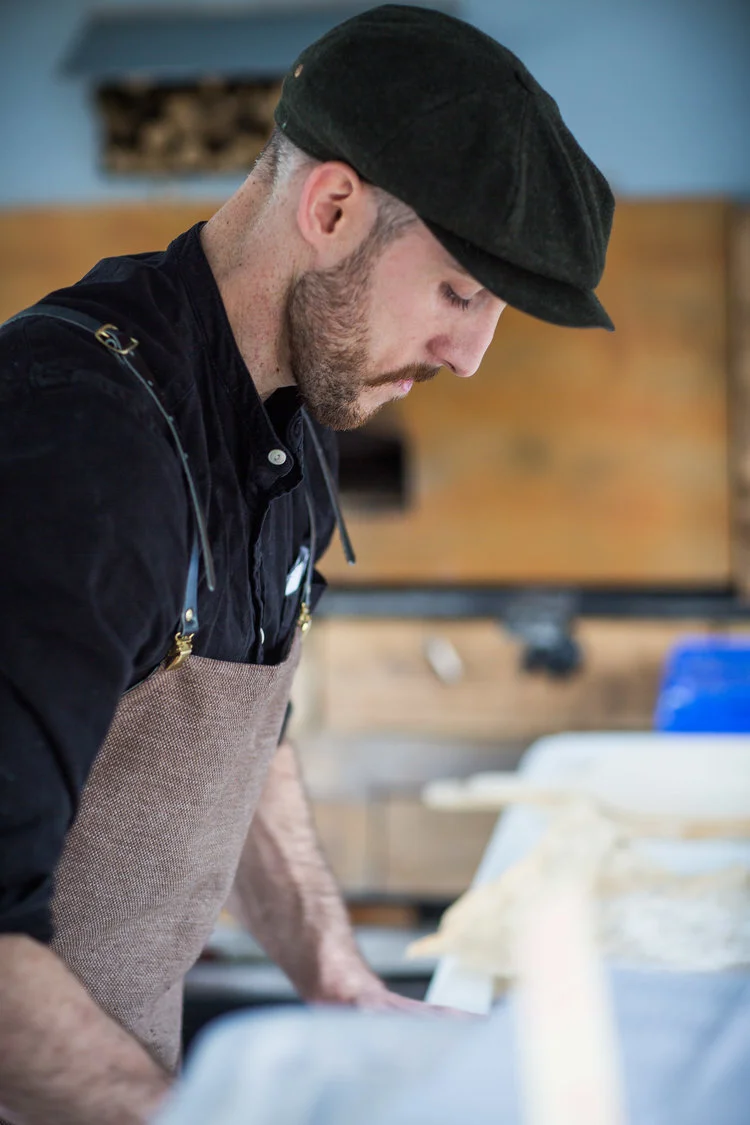 A man wearing a flat cap and apron working in a kitchen or workshop.
