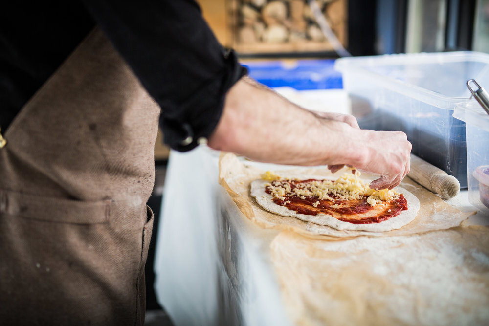 Person making pizza, adding cheese to dough with tomato sauce, wearing an apron in a kitchen setting.