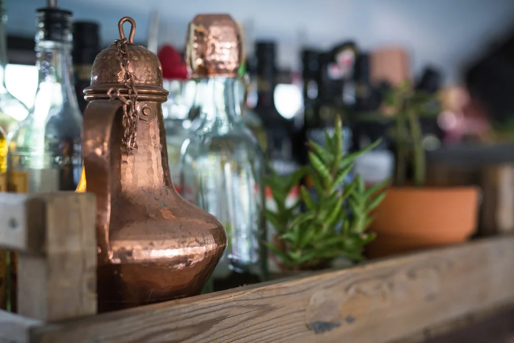 Copper kettle with chain, glass bottles, and small potted plant on wooden shelf.