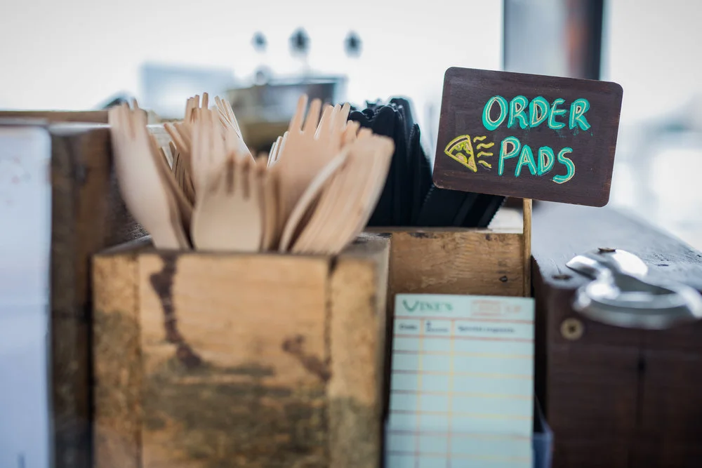 Wooden containers holding disposable forks and order pads on a counter, with a small sign reading 'Order Pads'. The scene suggests a self-serve area in a café or restaurant.