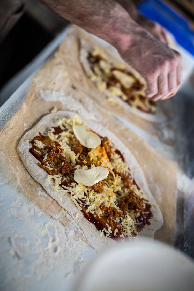 Person preparing pizza dough with toppings including cheese and meat on a floured surface.