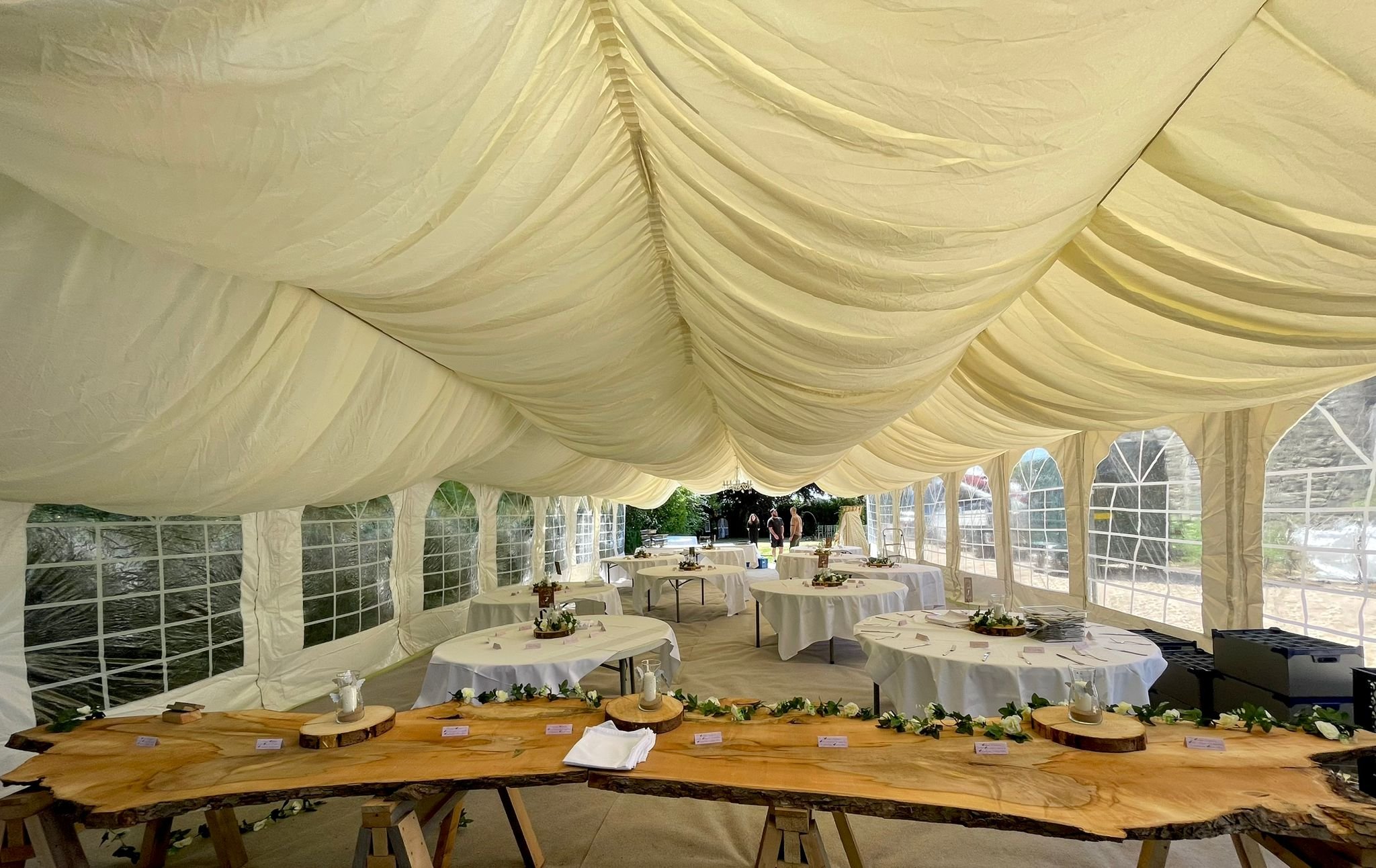 Interior of a decorated wedding marquee with white draped ceiling, round tables covered in white tablecloths, and a rustic wooden table with greenery and small signs.