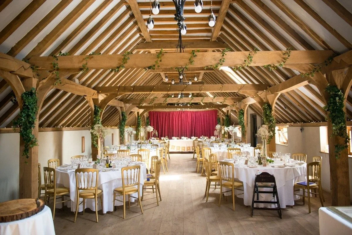 Event space decorated with round tables covered in white tablecloths, gold chairs, floral centerpieces, and a stage with a red curtain, in a rustic wooden building with exposed beams and greenery.