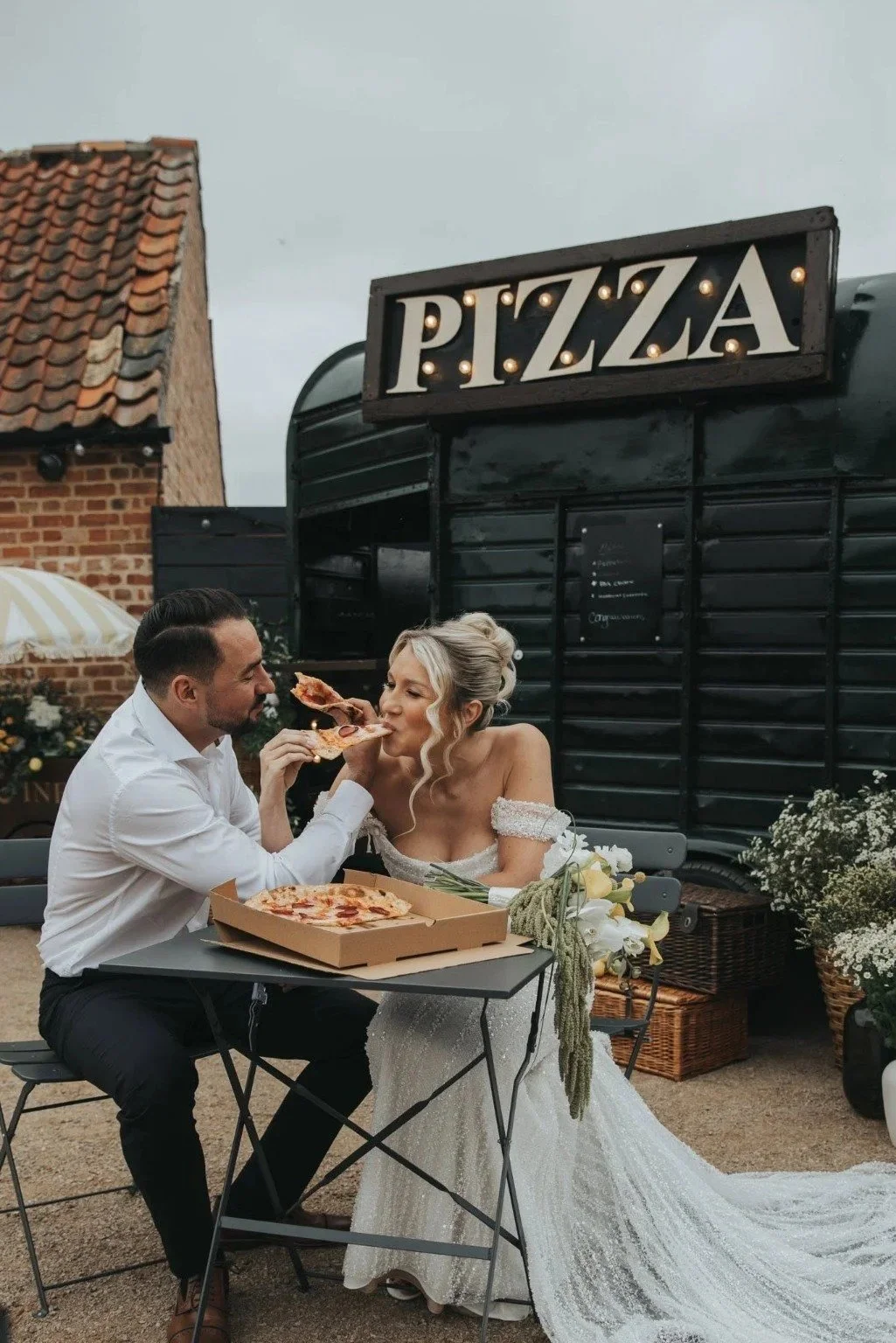 A man and woman, dressed in wedding attire, sharing a slice of pizza at an outdoor table with a pizza box, in front of a black pizza stand with a sign. The woman is wearing a white wedding gown with lace details, and the man is wearing a white shirt and black pants.