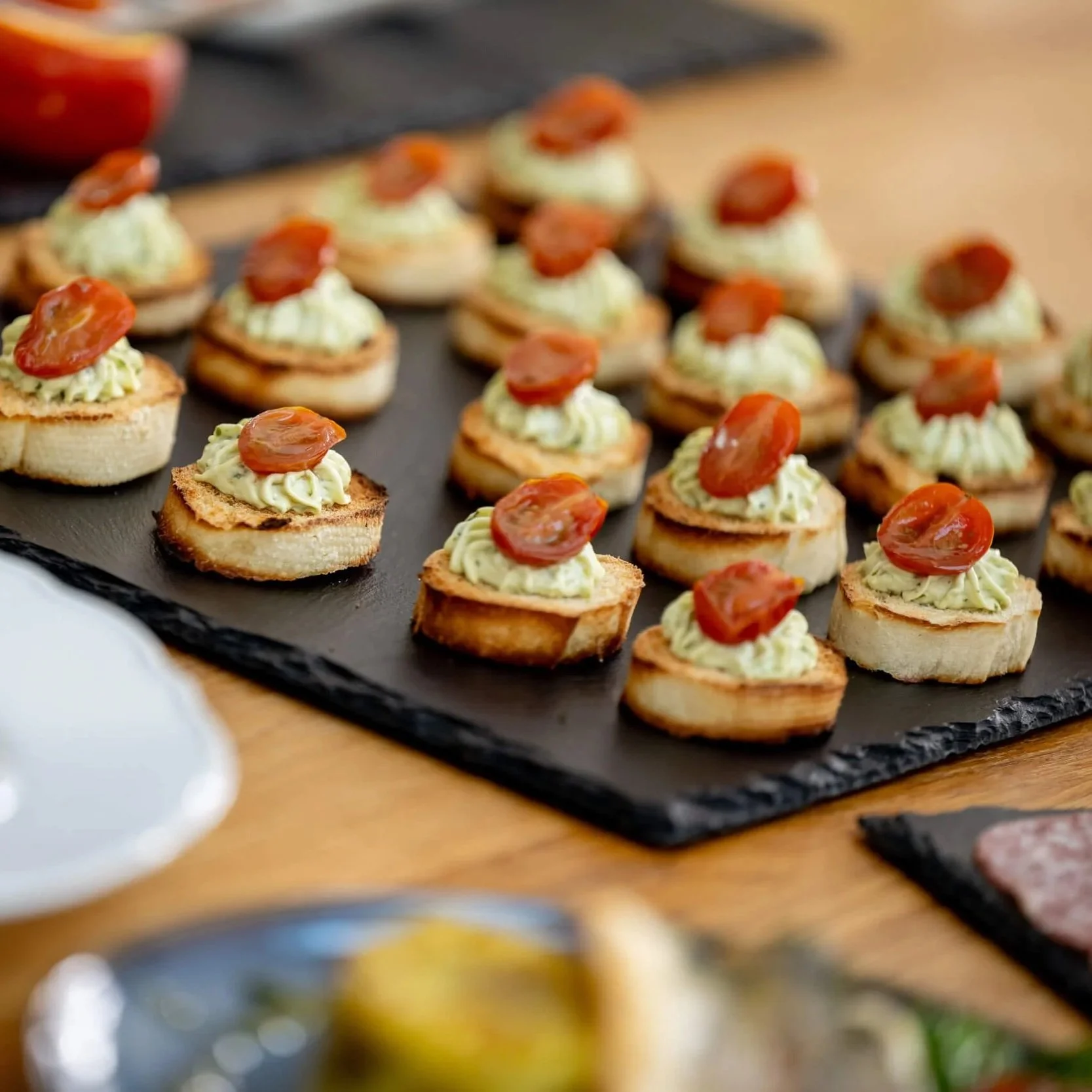 Assorted crostinis topped with cream cheese and cherry tomato slices on a black slate serving tray.