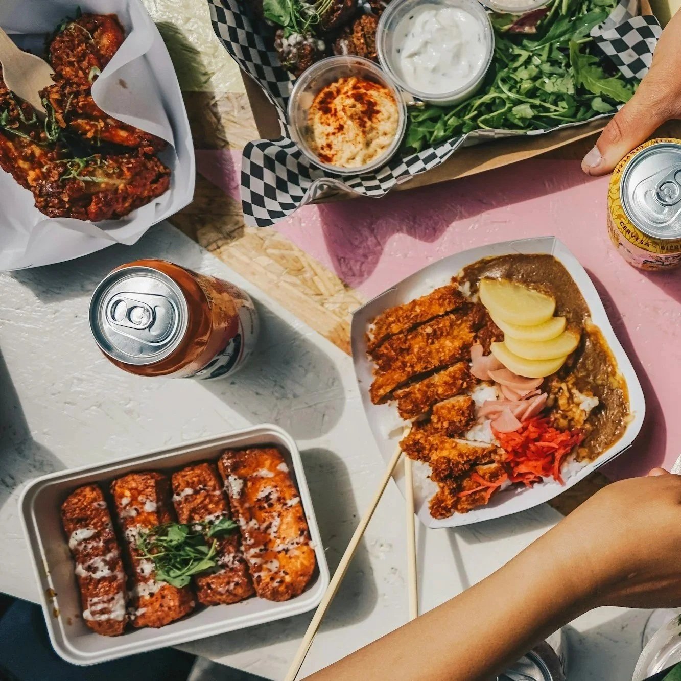 A table with a variety of Asian dishes, including fried chicken, sushi, curry with rice and pickled vegetables, and a tray of fried chicken wings with dipping sauces, along with two cans of soda, some green leafy vegetables, and bowls of white sauce and casserole.