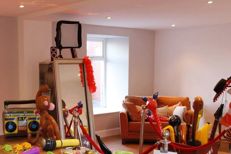 Room with a variety of party props, including inflatable animals, a retro boombox, a mirror with a red feather boa, and a couch in the background. Bright lighting enhances the playful atmosphere.