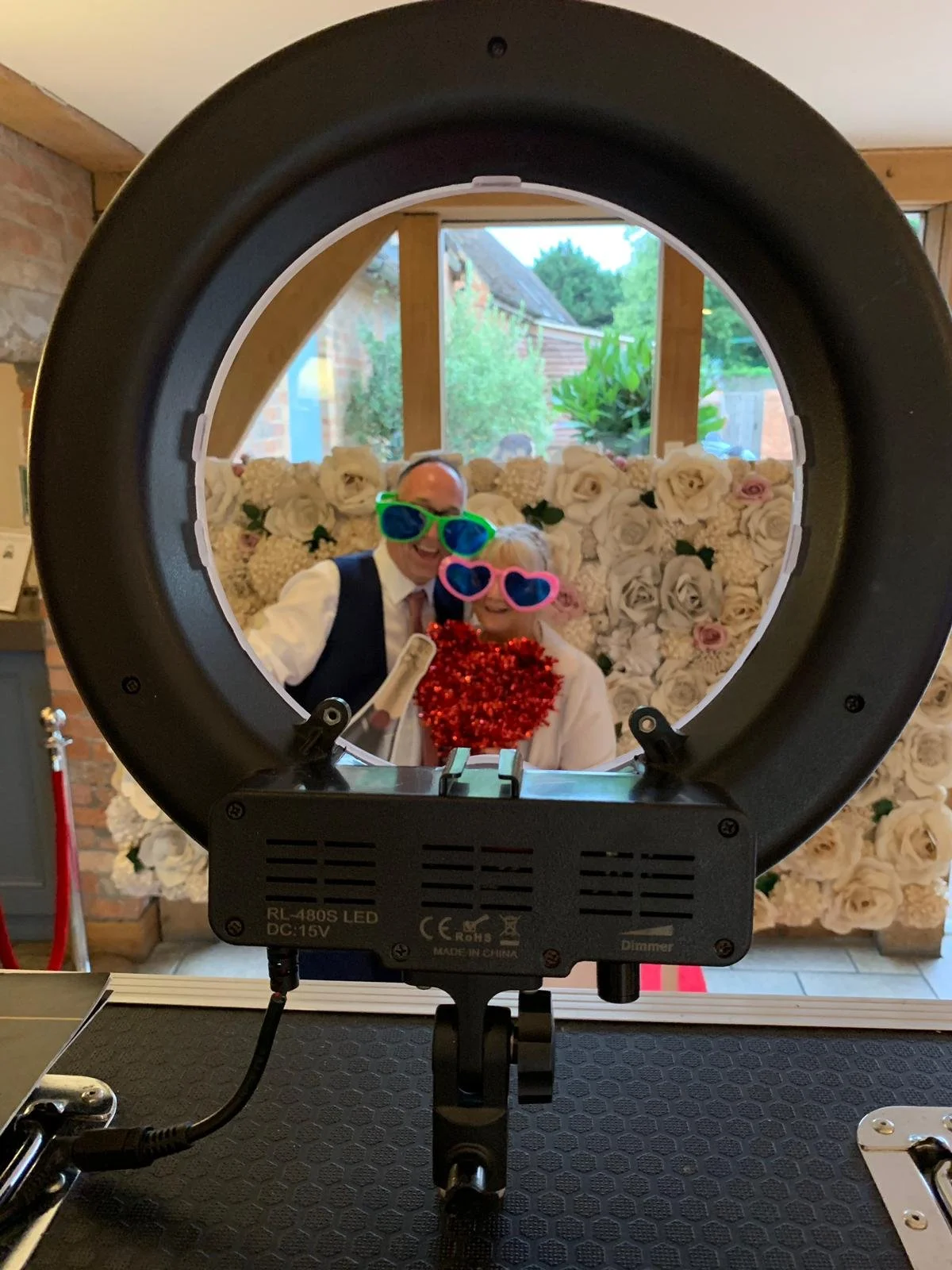 A couple posing for a photo behind a ring light, wearing colorful oversized glasses and a red tinsel boa. The background features a floral wall with white roses.