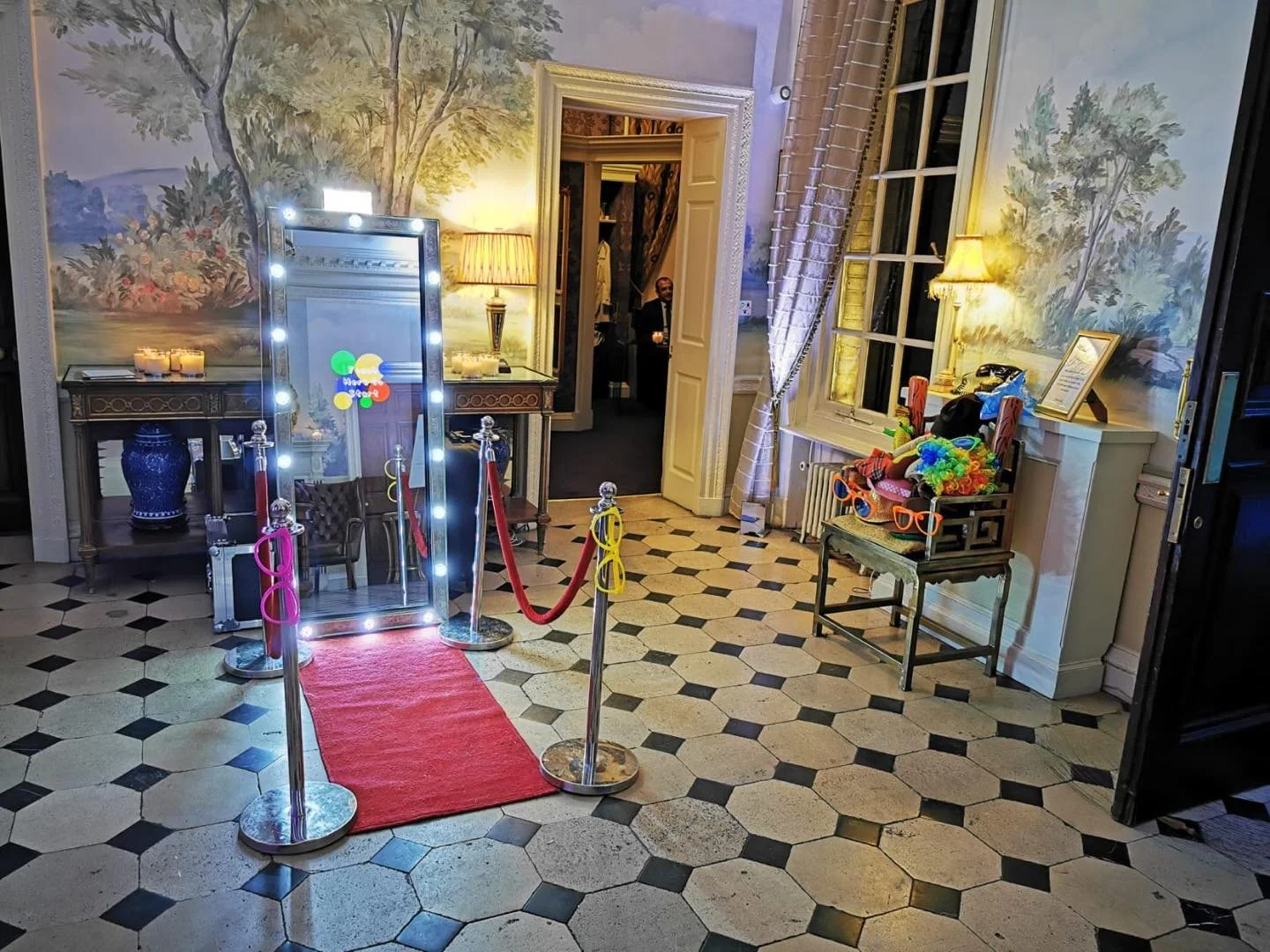 Elegant room setup with a large mirror decorated with lights, a red carpet, and stanchions. Colorful props and novelty sunglasses are displayed on a chair. The room features a scenic mural, classic furniture, and warm lighting.