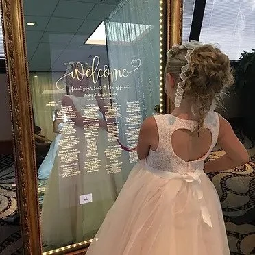 Young girl in white dress looking at a decorative mirror with seating chart and welcome message.