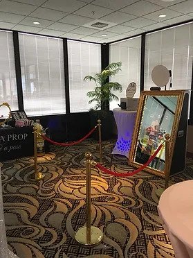 Photo booth setup with mirror, red rope barriers, and decorative lighting in a room with large windows and patterned carpet.