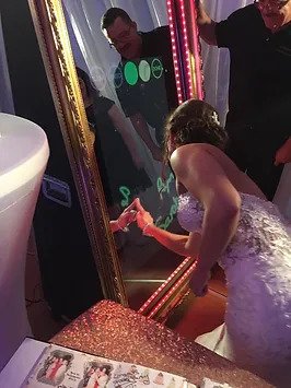 A bride in a wedding dress interacts with a touch screen mirror photo booth. The mirror is framed in gold and surrounded by lights. A piece of paper with photo prints is on a nearby table.