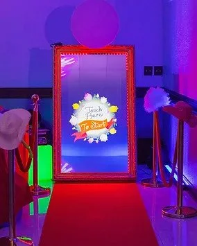 Interactive mirror photo booth with touch screen, surrounded by decorations and red carpet.