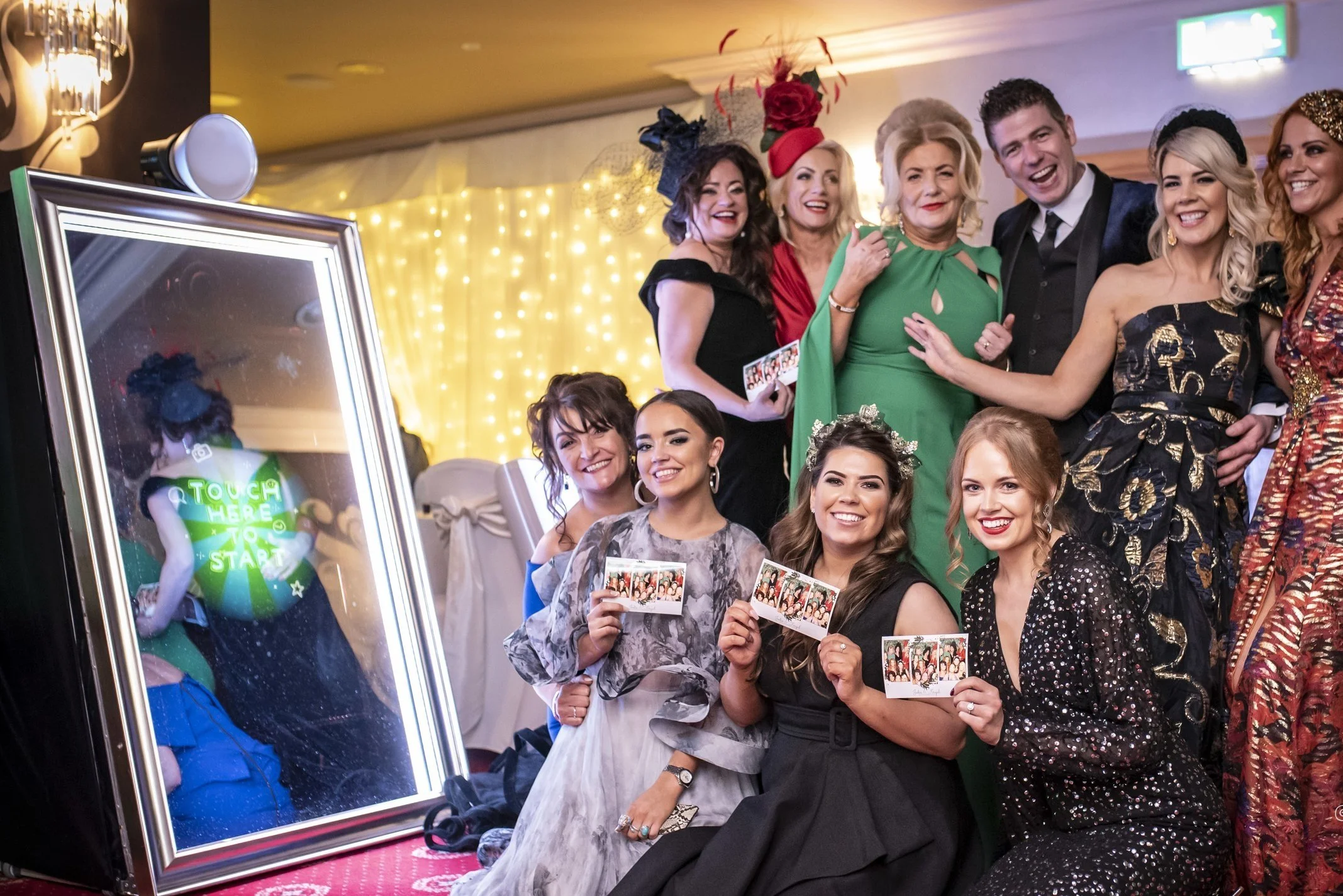 Group of people dressed in formal attire smiling and holding photo strips at a party, next to a digital photo booth mirror.
