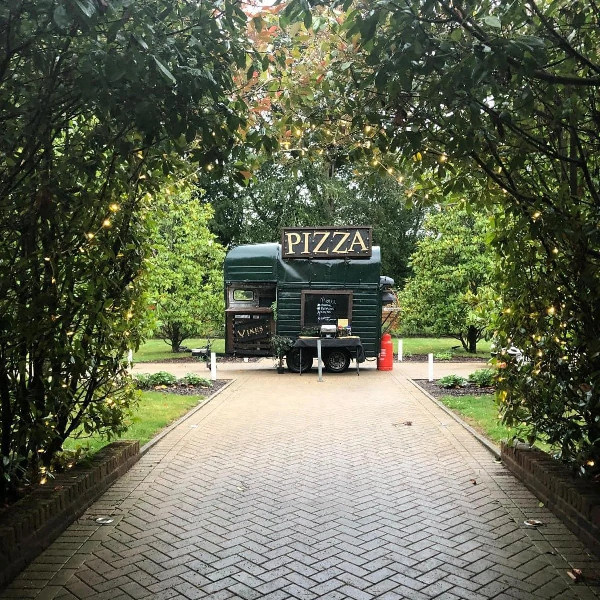 Mobile pizza truck on a paved path surrounded by greenery and small lights.