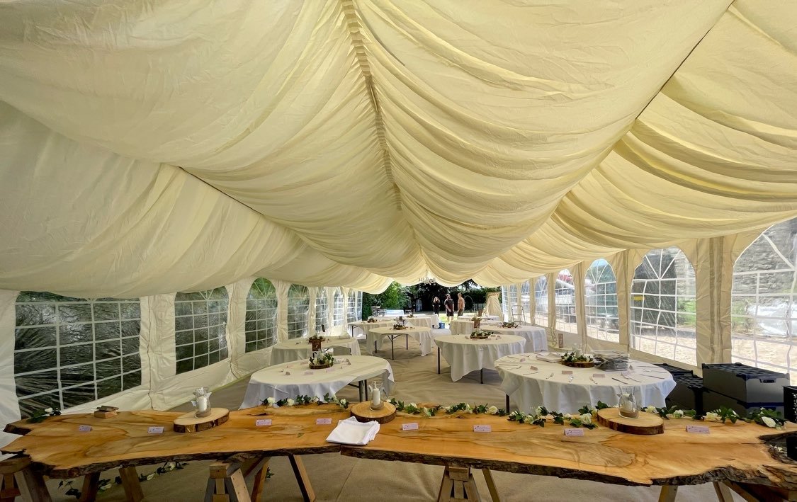 Inside a decorated event tent with draped fabric ceiling, round tables with white tablecloths, and a wooden serving table with floral arrangements.