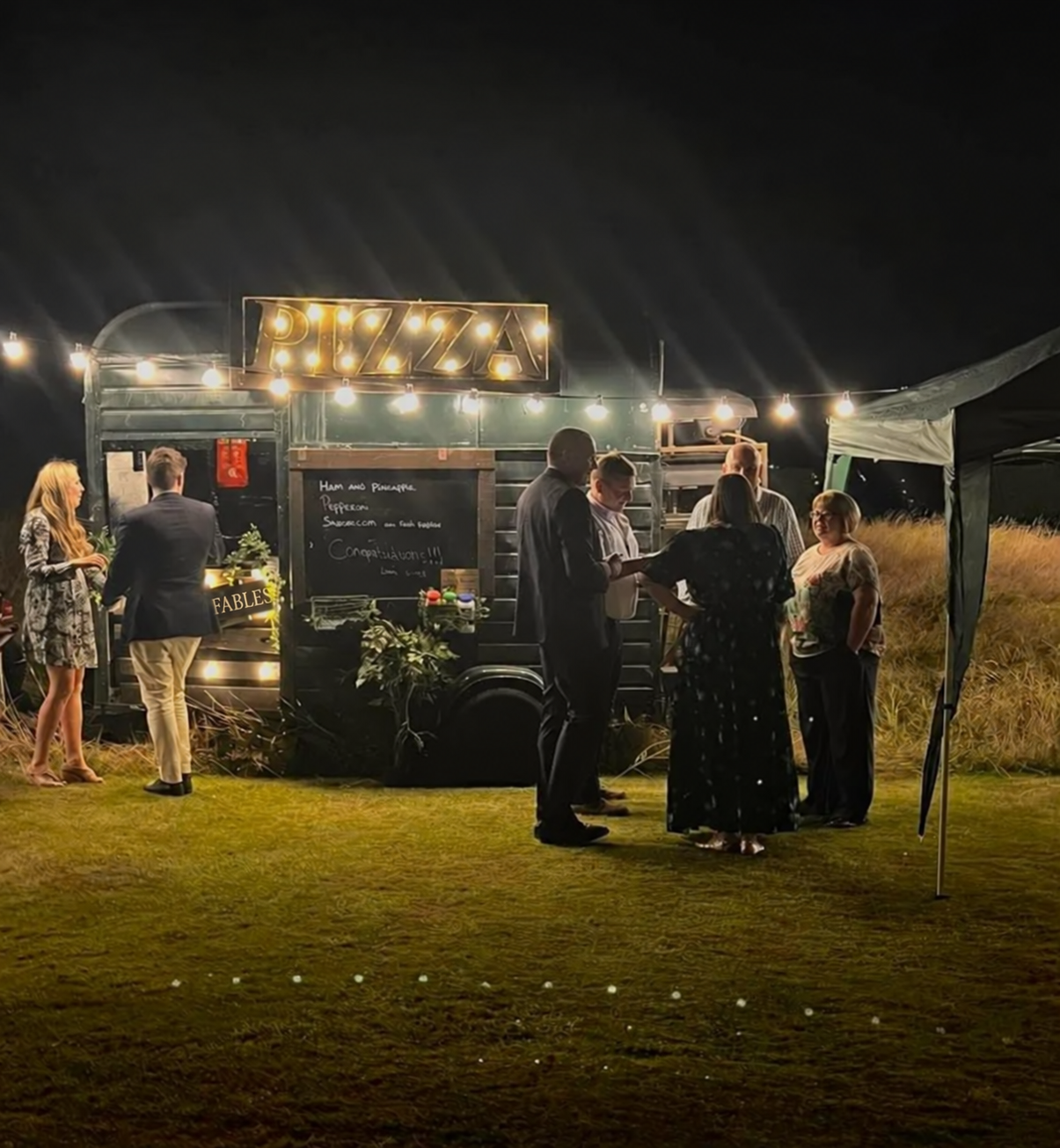 Guests gathered beside an illuminated Fables pizza horsebox at an evening event