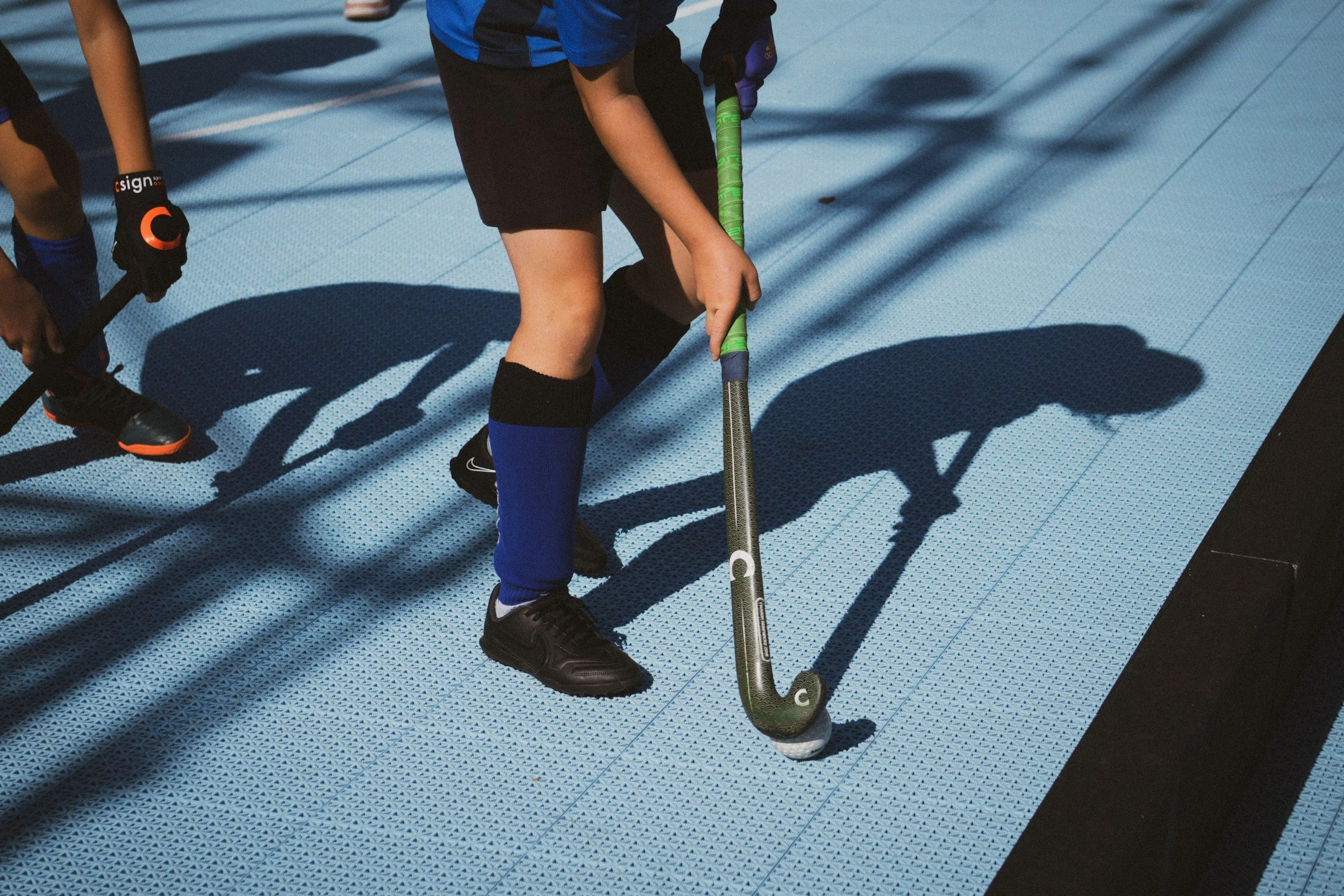 Children playing street hockey on a blue textured surface, with one child in black shoes and black shorts holding a hockey stick, and another child partially visible on the left.