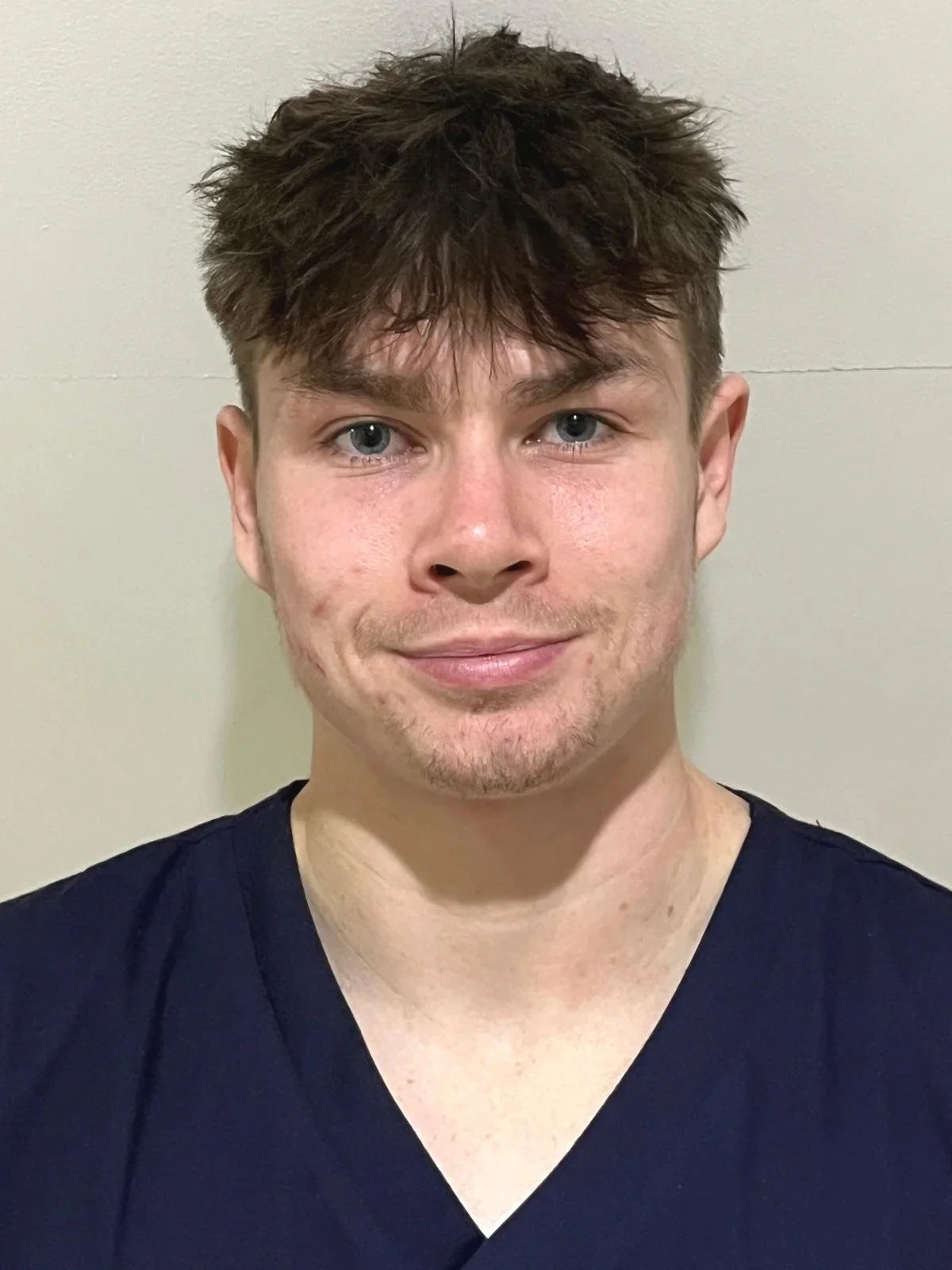 A young man with light skin, blue eyes, and short, tousled brown hair. He is smiling slightly and is wearing a dark blue V-neck shirt. The background is a plain, off-white wall.