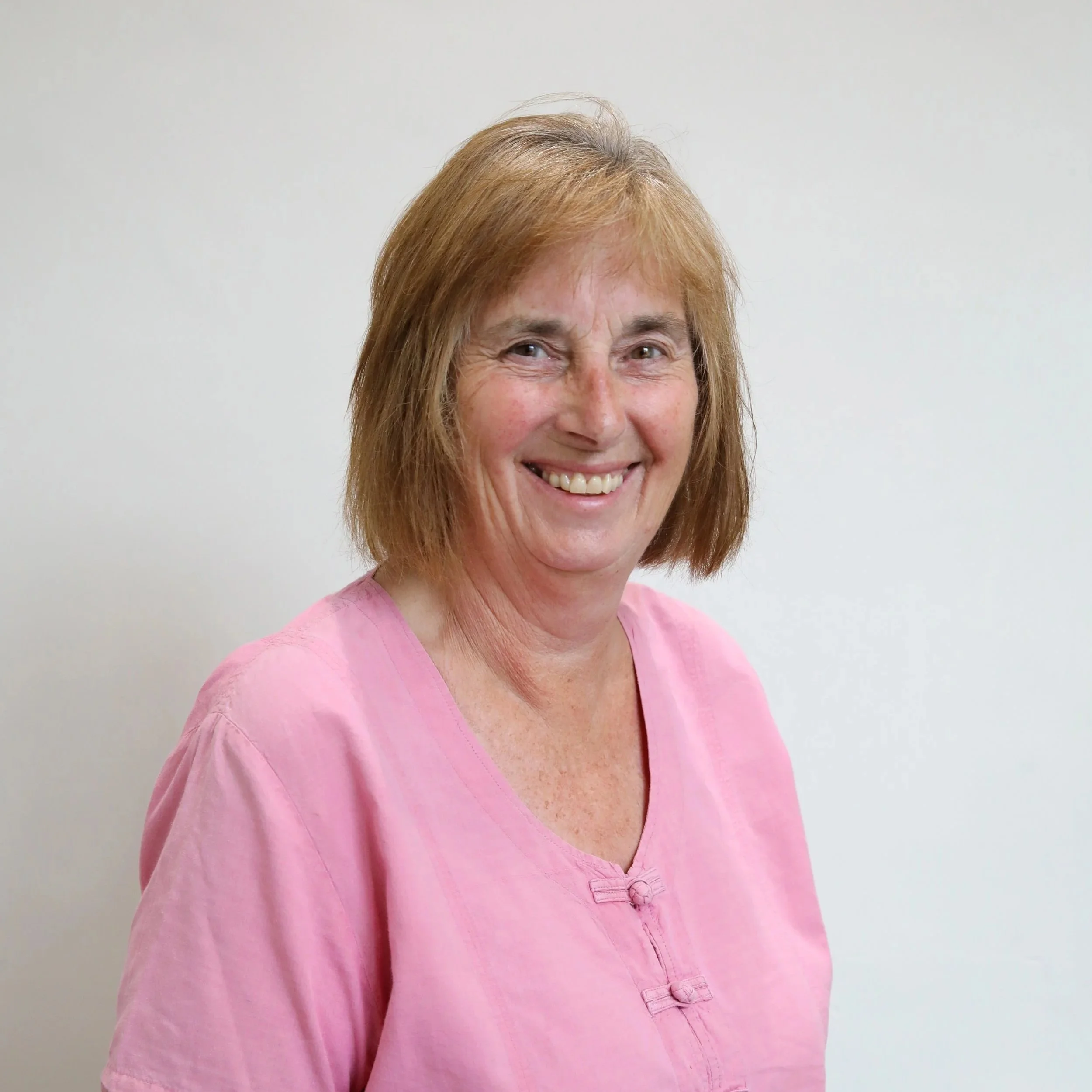 Close-up of a woman with shoulder-length reddish-brown hair, smiling and wearing a pink top, standing against a plain white background.