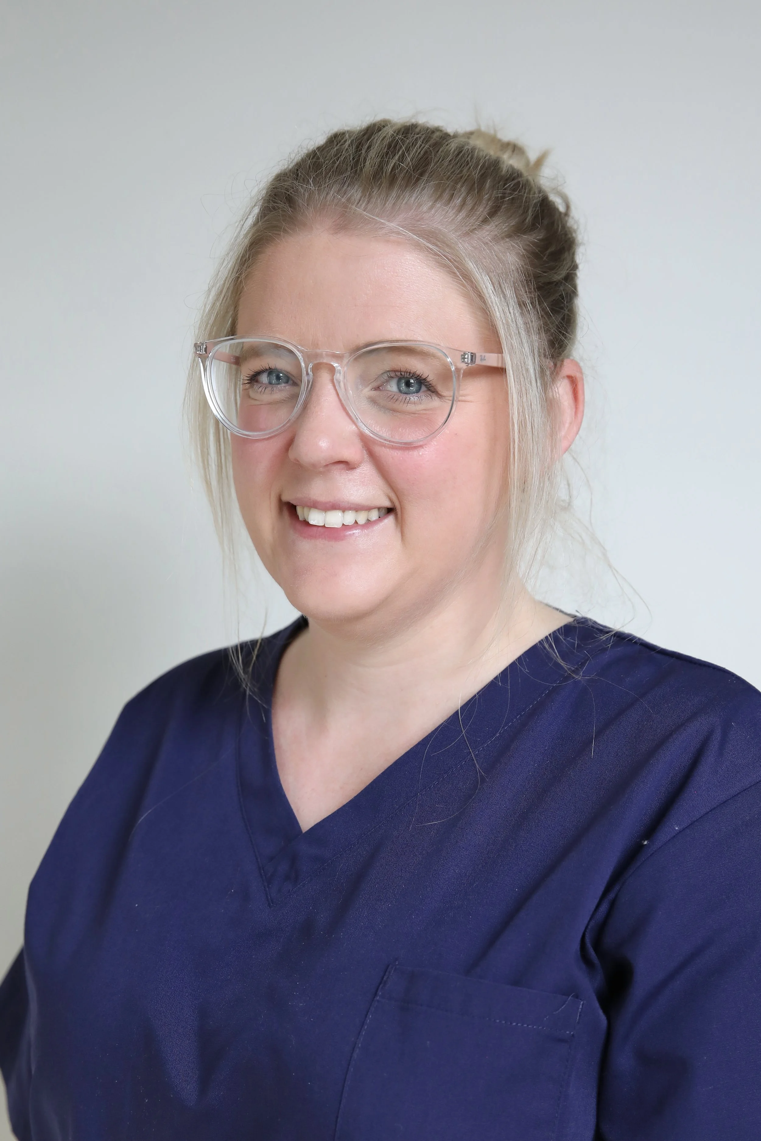 A woman with blonde hair tied back, wearing clear glasses and navy blue medical scrubs, smiling against a plain white background.