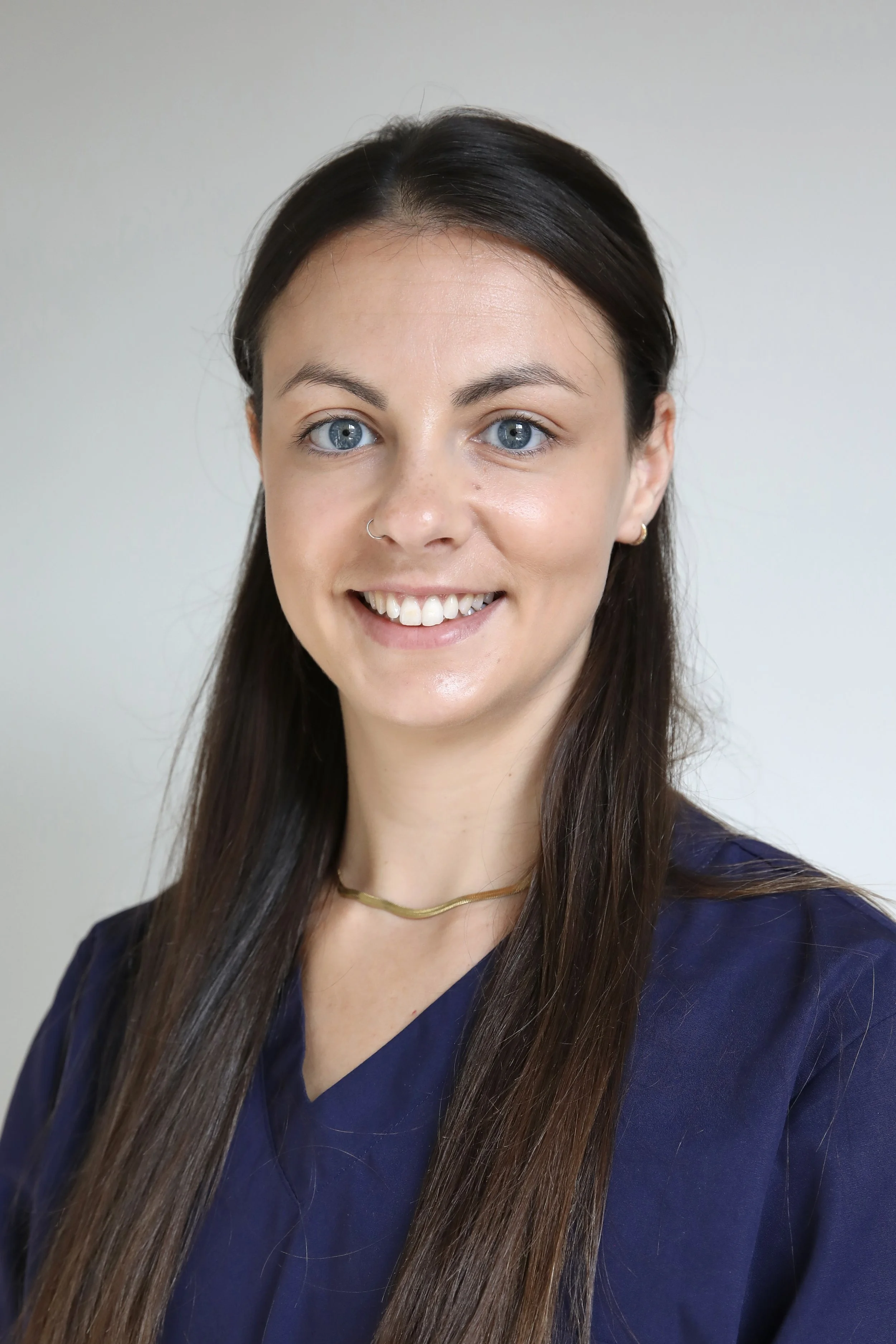 A young woman with long brown hair, light skin, and blue eyes, smiling, wearing a navy blue top, a gold necklace, standing against a plain light-colored background.