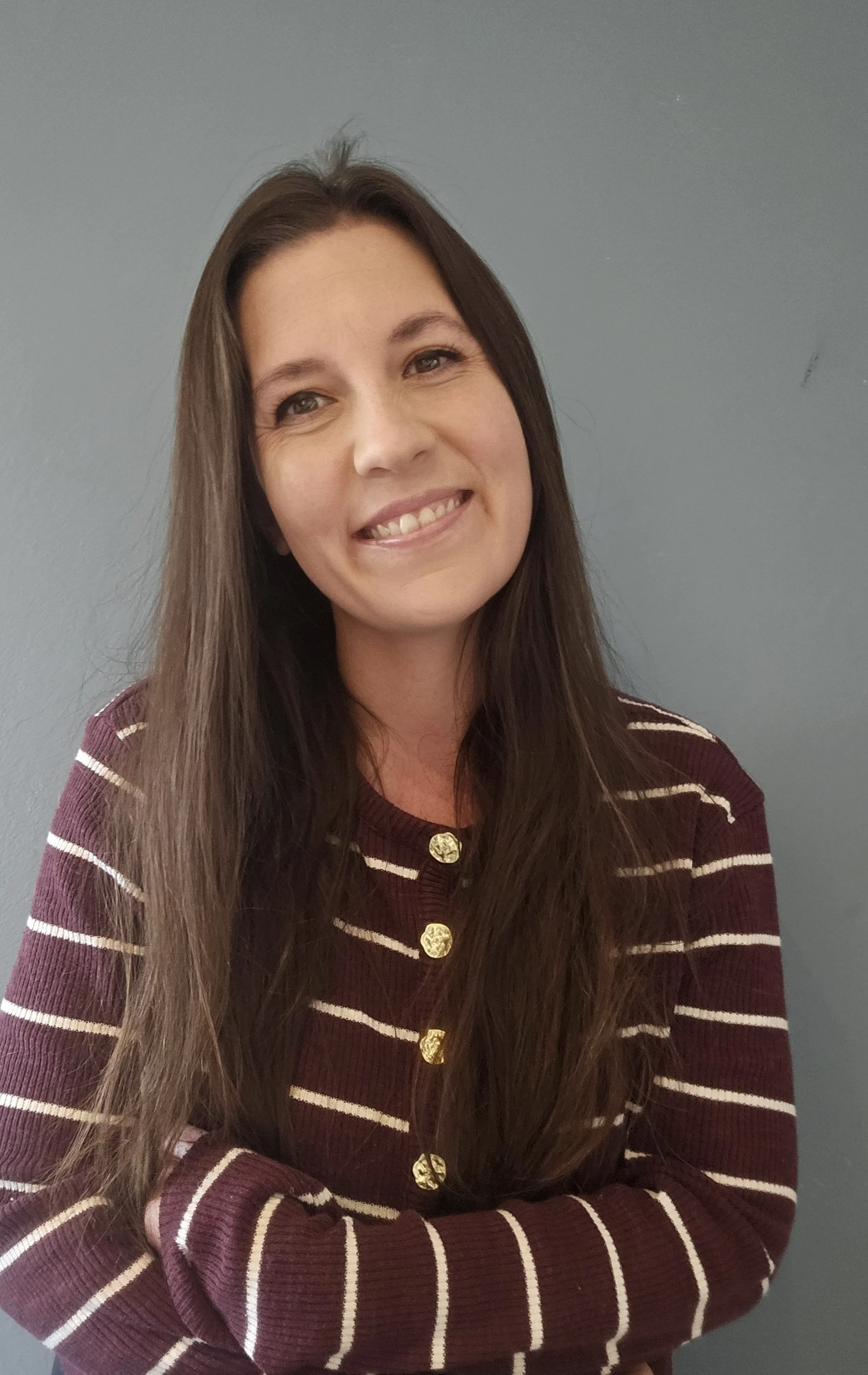 A woman with long brown hair smiling, arms crossed, wearing a maroon and white striped cardigan with gold buttons, standing against a gray wall.