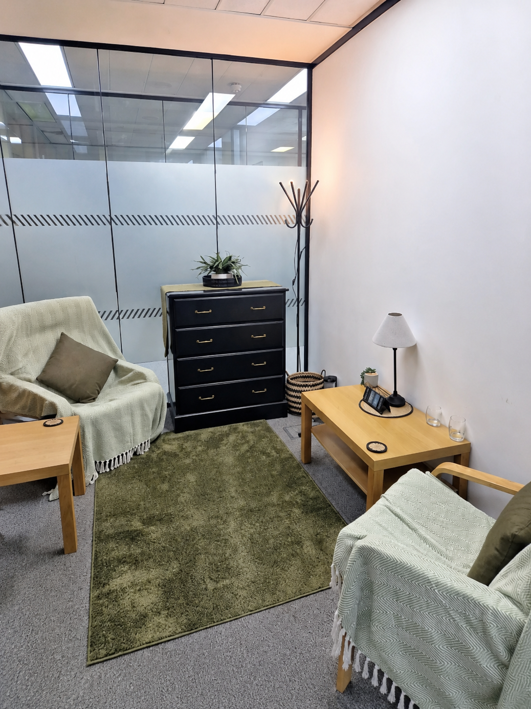 Office waiting area with two beige armchairs with throw blankets, a small wooden coffee table with glasses, a black dresser with a potted plant, a coat rack, a side table with a lamp, phone, and plants, and a green rug on gray carpet, separated by glass walls.