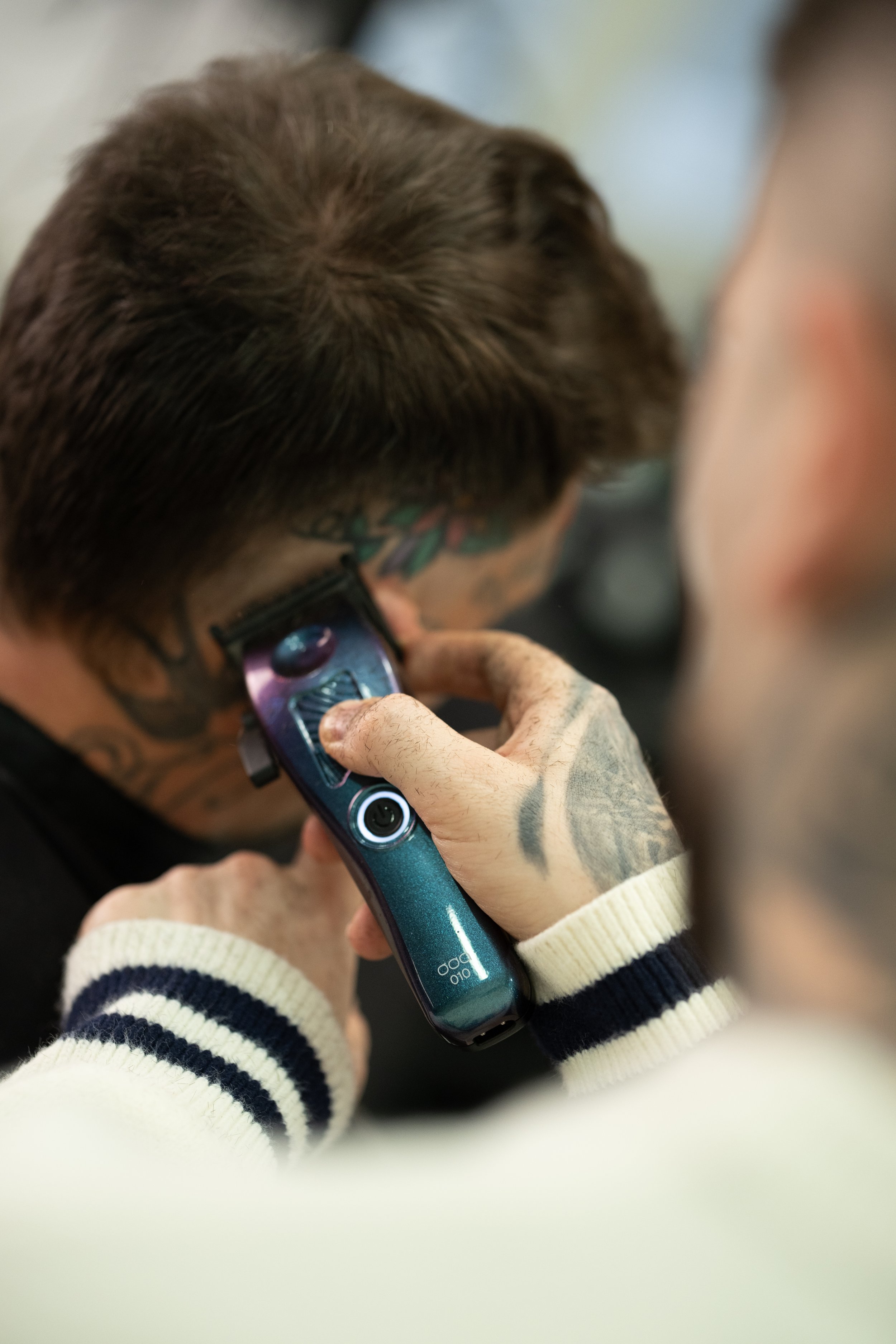 A person with tattoos and tattoos on their neck is getting a haircut using hair clippers. The person is wearing a striped sweater.