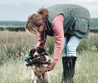 A woman in outdoor attire bends down to hold a dog by a lake or river in a grassy field.