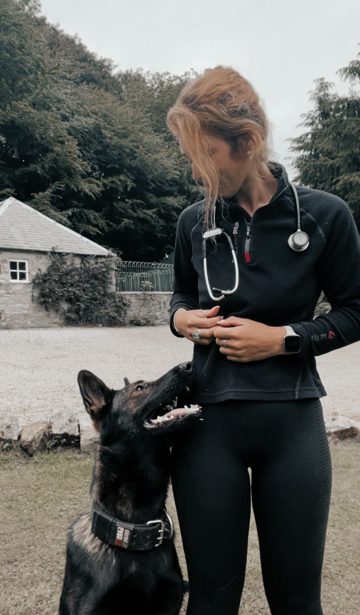 A woman with blonde hair in athletic clothing standing outdoors with a German Shepherd dog, which is looking up at her and wearing a collar. The background features trees and a small building.