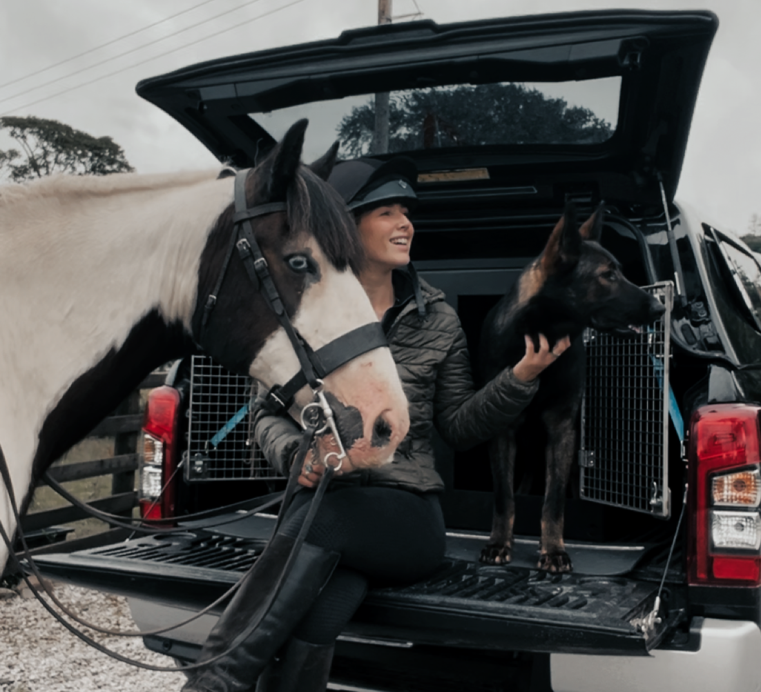 A woman kneeling in the back of a truck with a white and black horse and a black and tan dog, all looking to the right.