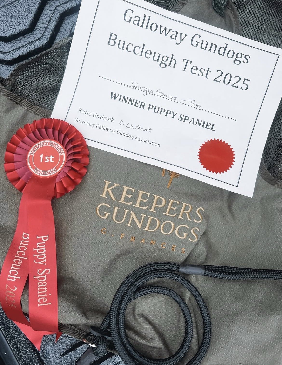 A Galloway Gundogs award certificate and red ribbon rosette with the text '1st Galloway Gundog Association' on it. The certificate recognizes a winning puppy spaniel, awarded at the 2025 Bouclough Test, with the breeder's name, Katie Unthank. The ite