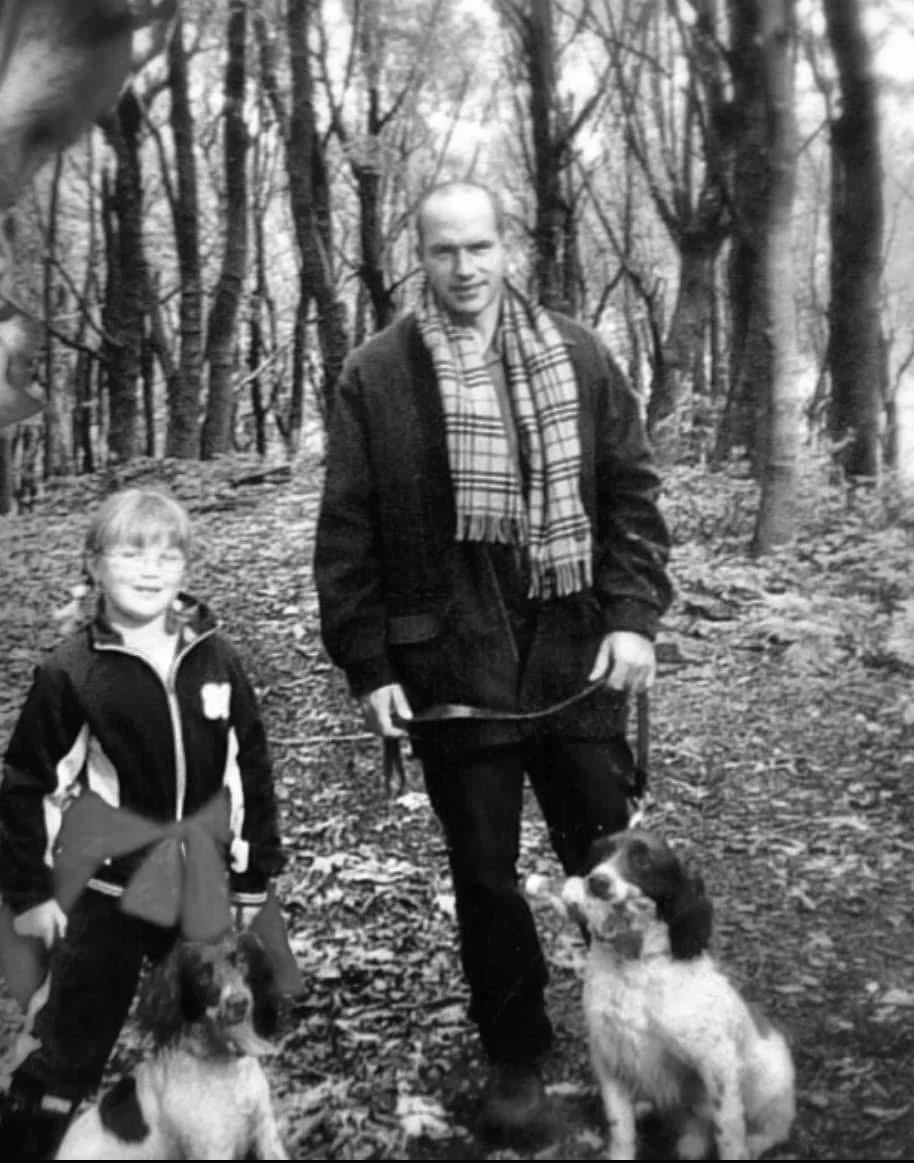 A black-and-white photo of a man and a young girl walking in a wooded area with two dogs. The man is holding a leash attached to one dog, and the girl is standing next to the other dog. The man is wearing a scarf and a jacket, and the girl is wearing a jacket with a bow. The trees are bare, suggesting late autumn or winter.