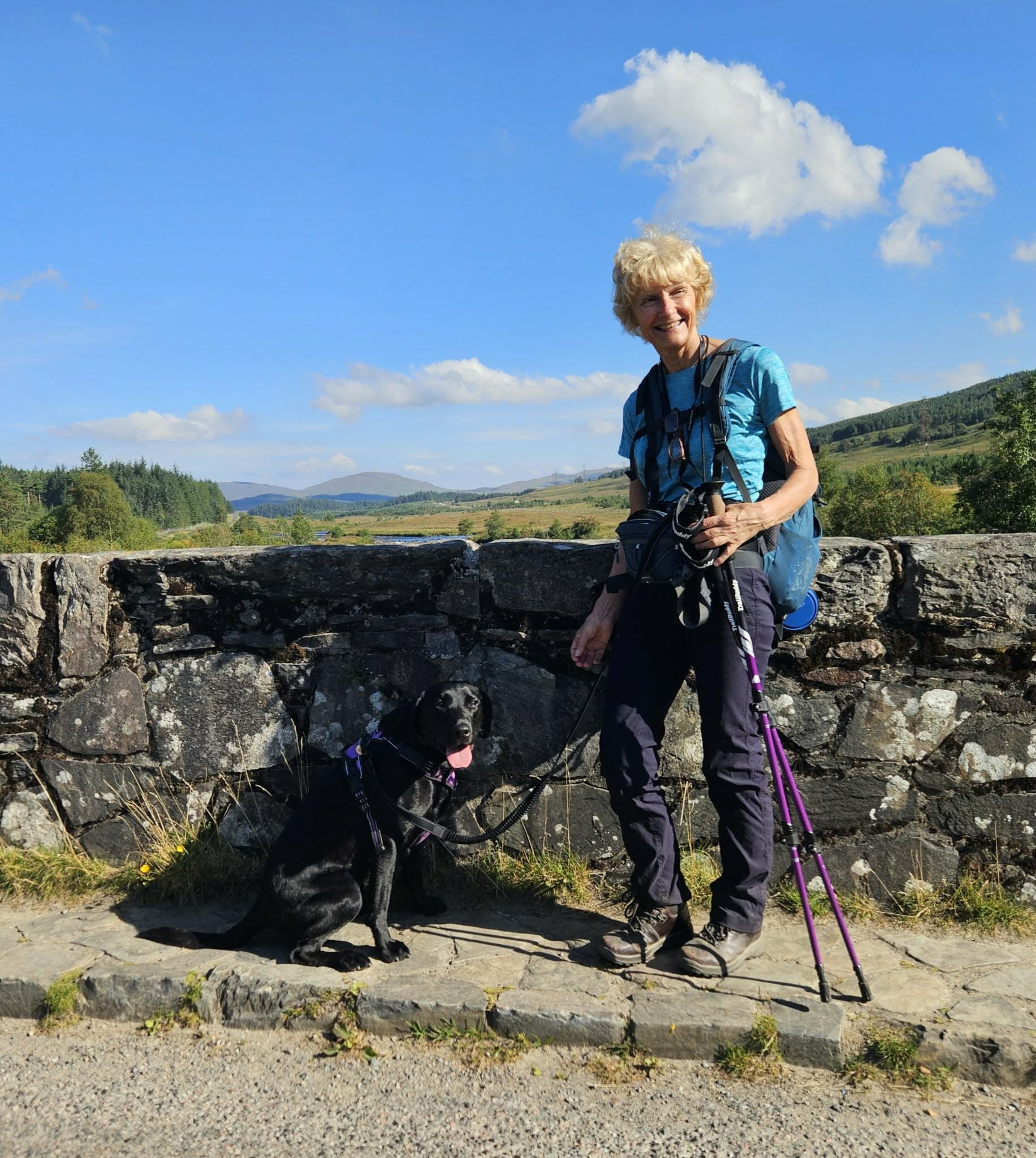 A woman with curly blonde hair smiling and standing outdoors with two black and white dogs on leashes, in front of a low stone wall, greenery, and a landscape with hills under a partly cloudy sky.