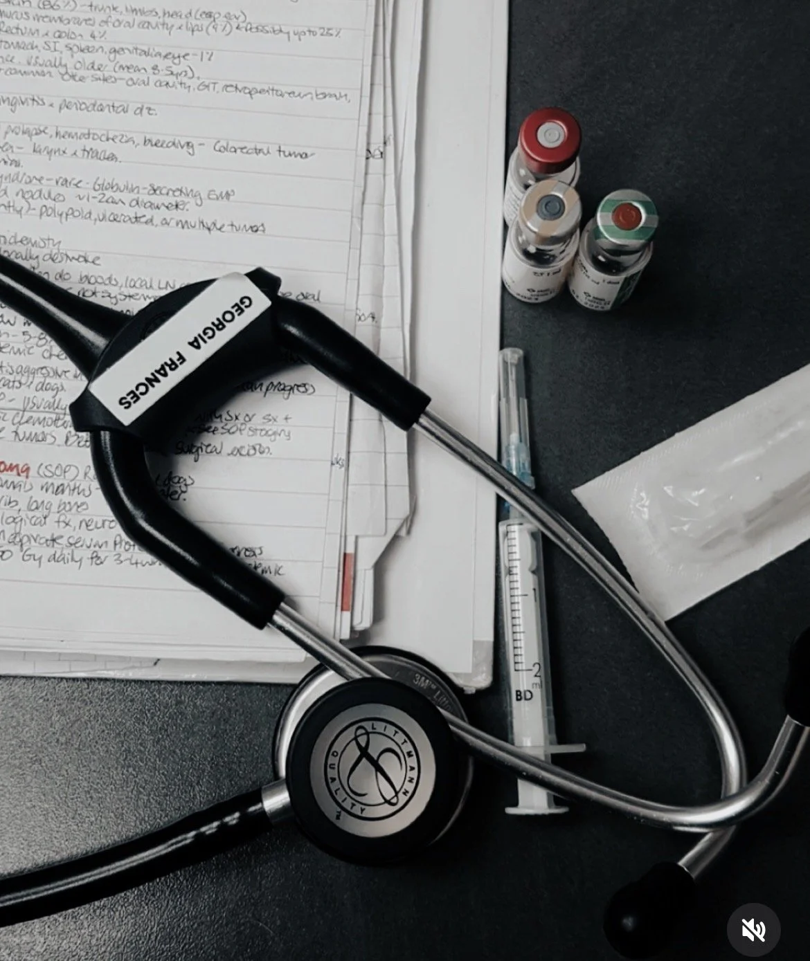 A stethoscope placed on medical notes, with medication bottles and a syringe nearby, on a dark surface.