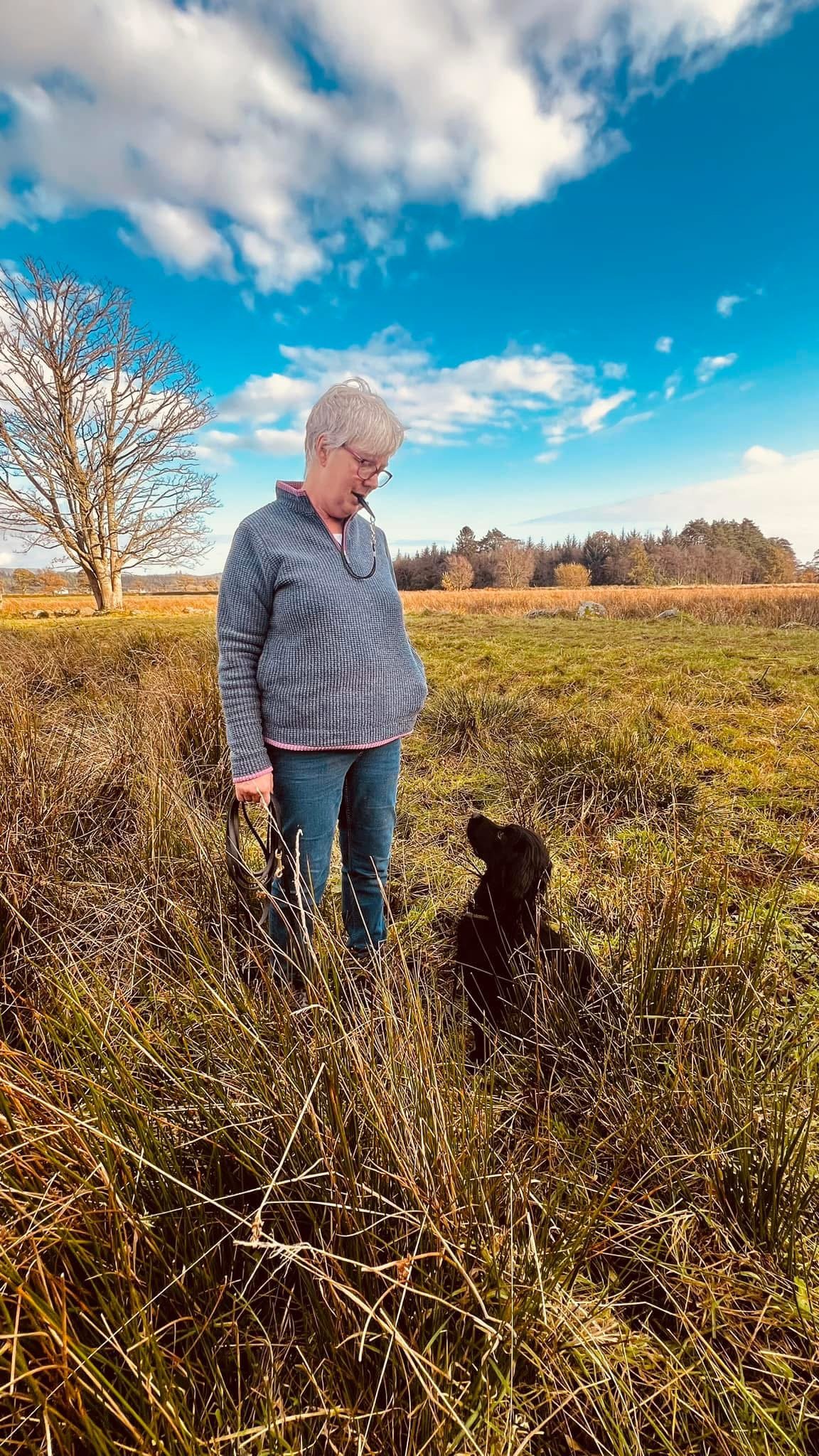A woman with short gray hair and glasses standing in a field of tall grass, looking down at a black dog sitting in front of her, under a partly cloudy sky.