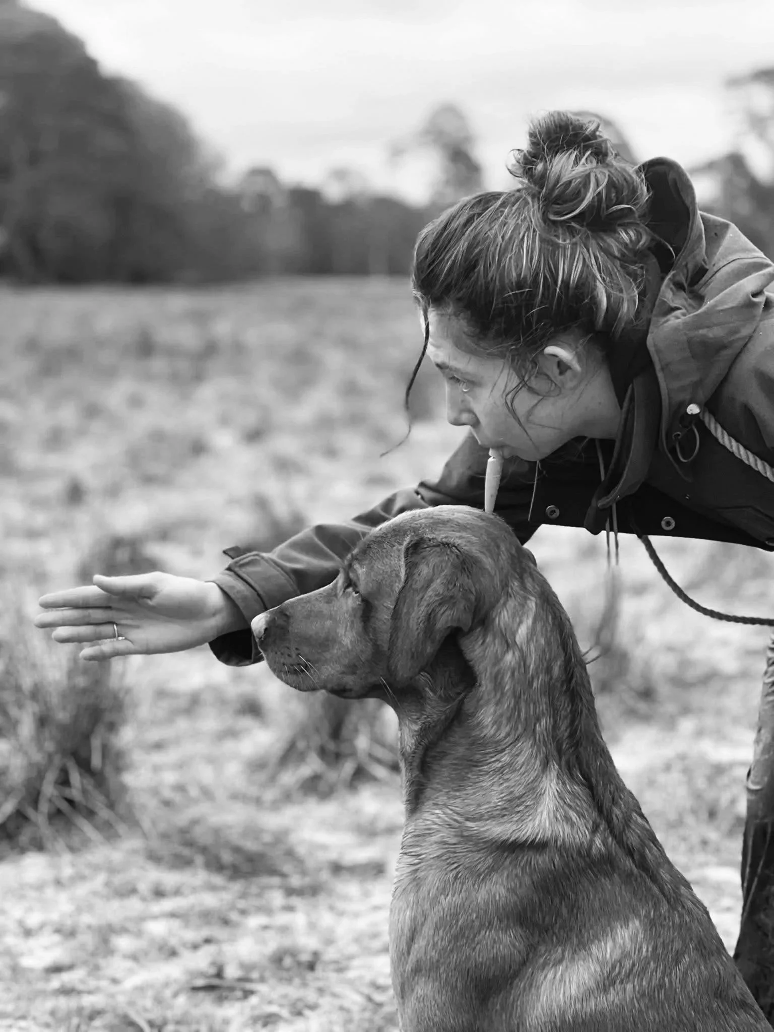 A woman with her hair in a bun is playing with a dog outdoors in black and white.