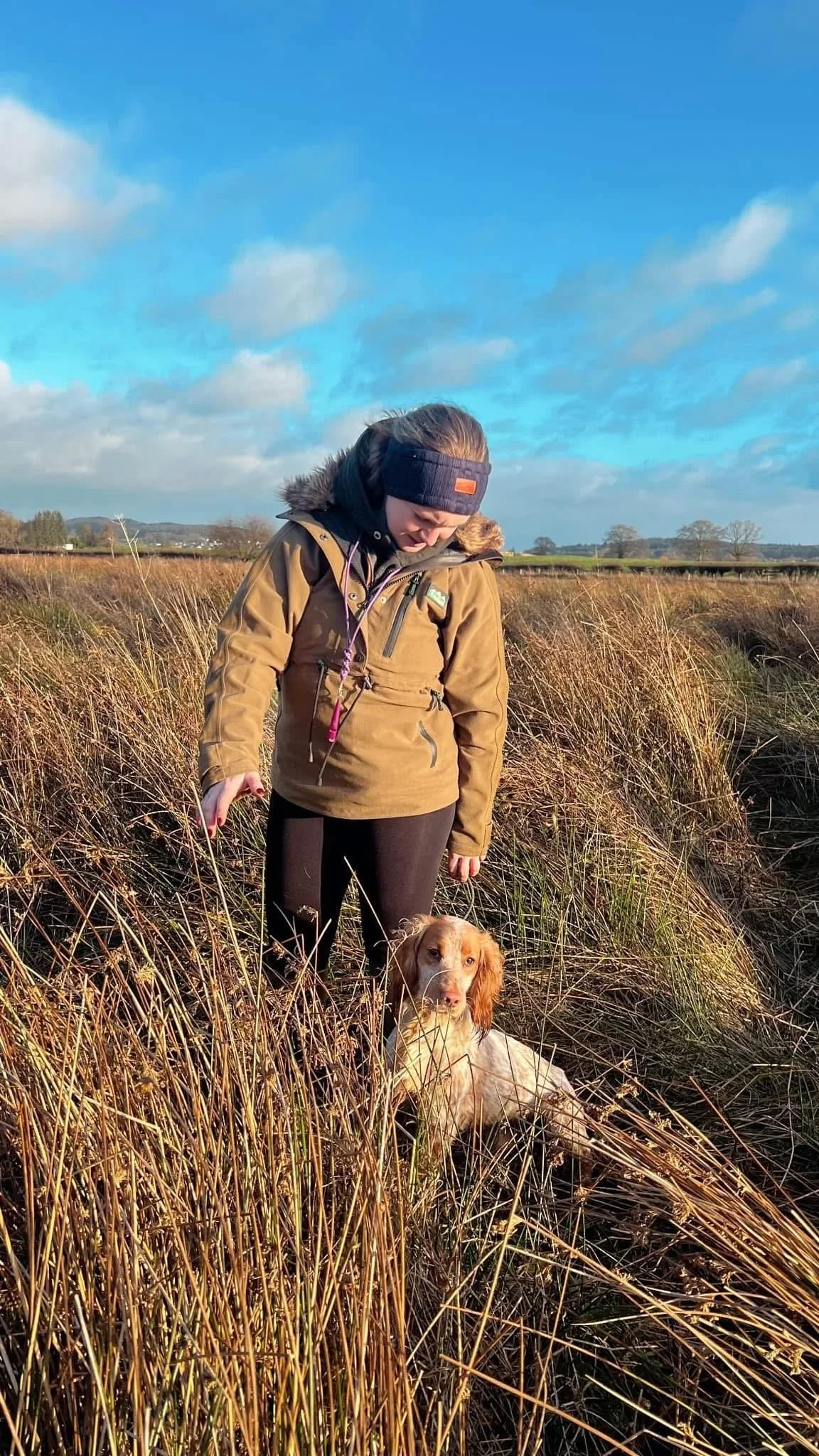 A woman with a dog in a grassy field on a sunny day with a partly cloudy sky.