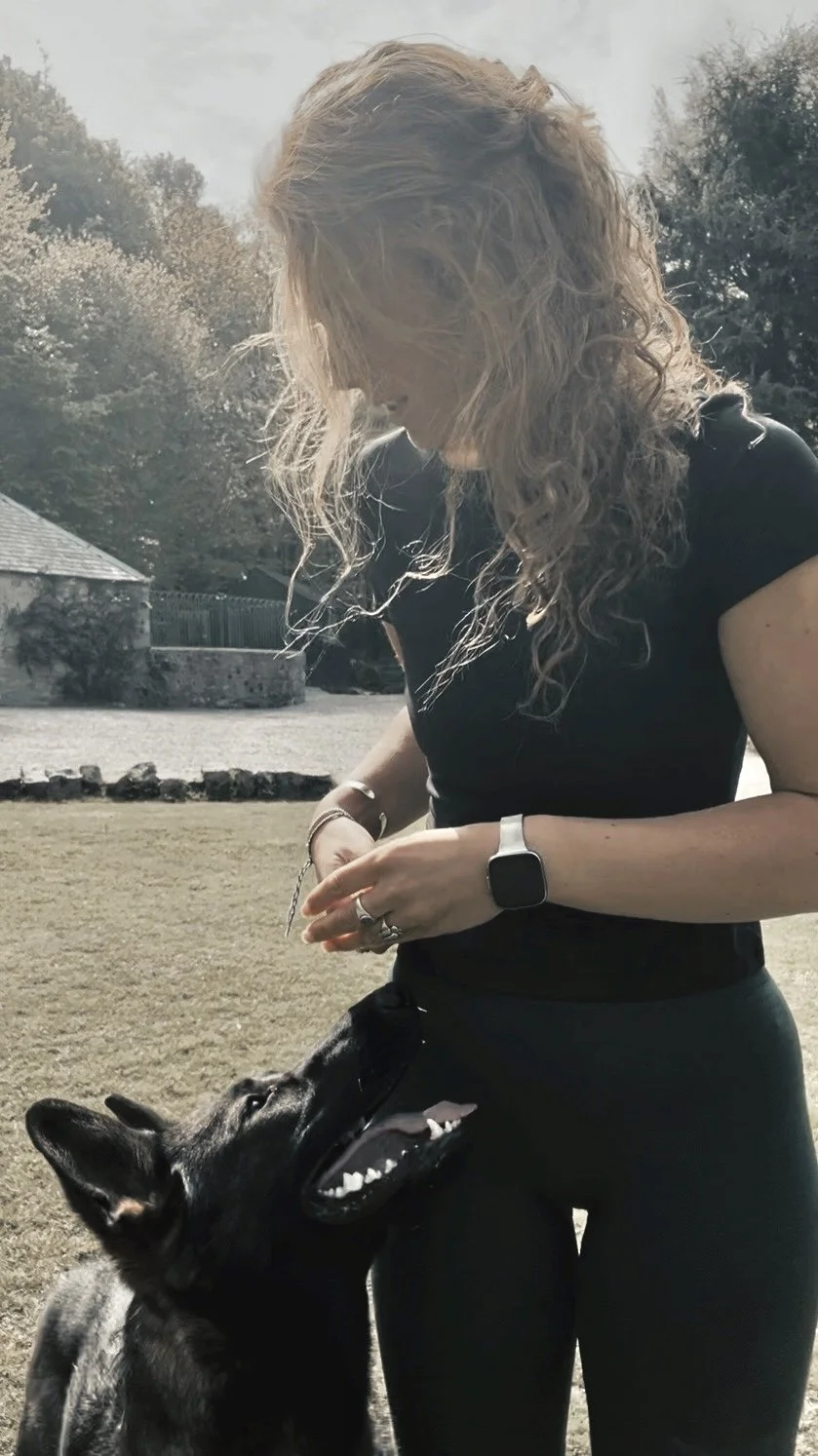 A woman with curly hair and a smartwatch stands outdoors with a black dog that is sitting and looking up at her, with trees and a building in the background.