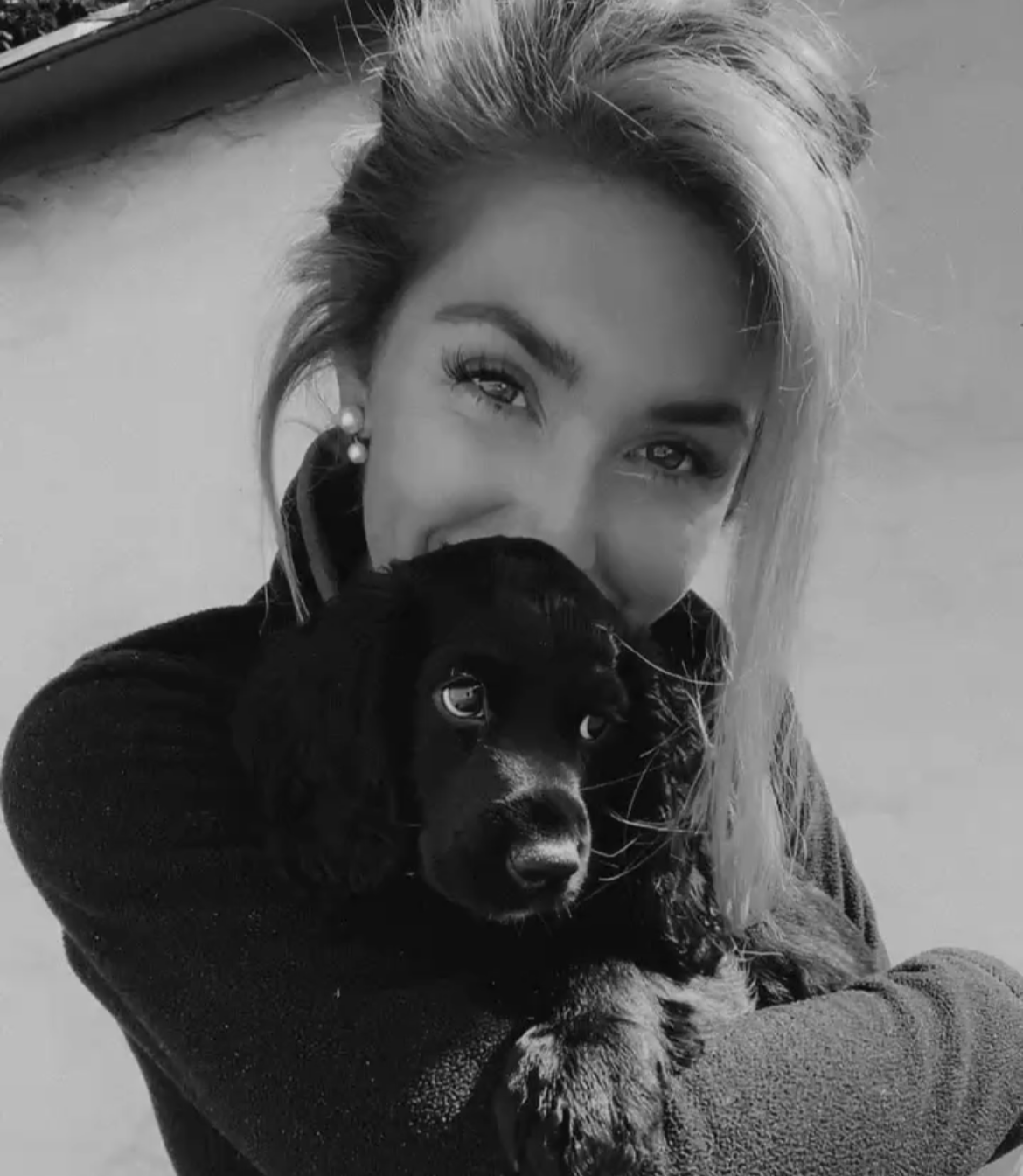 A woman with blonde hair and earrings holding a black puppy close to her face, both looking at the camera, in black and white.