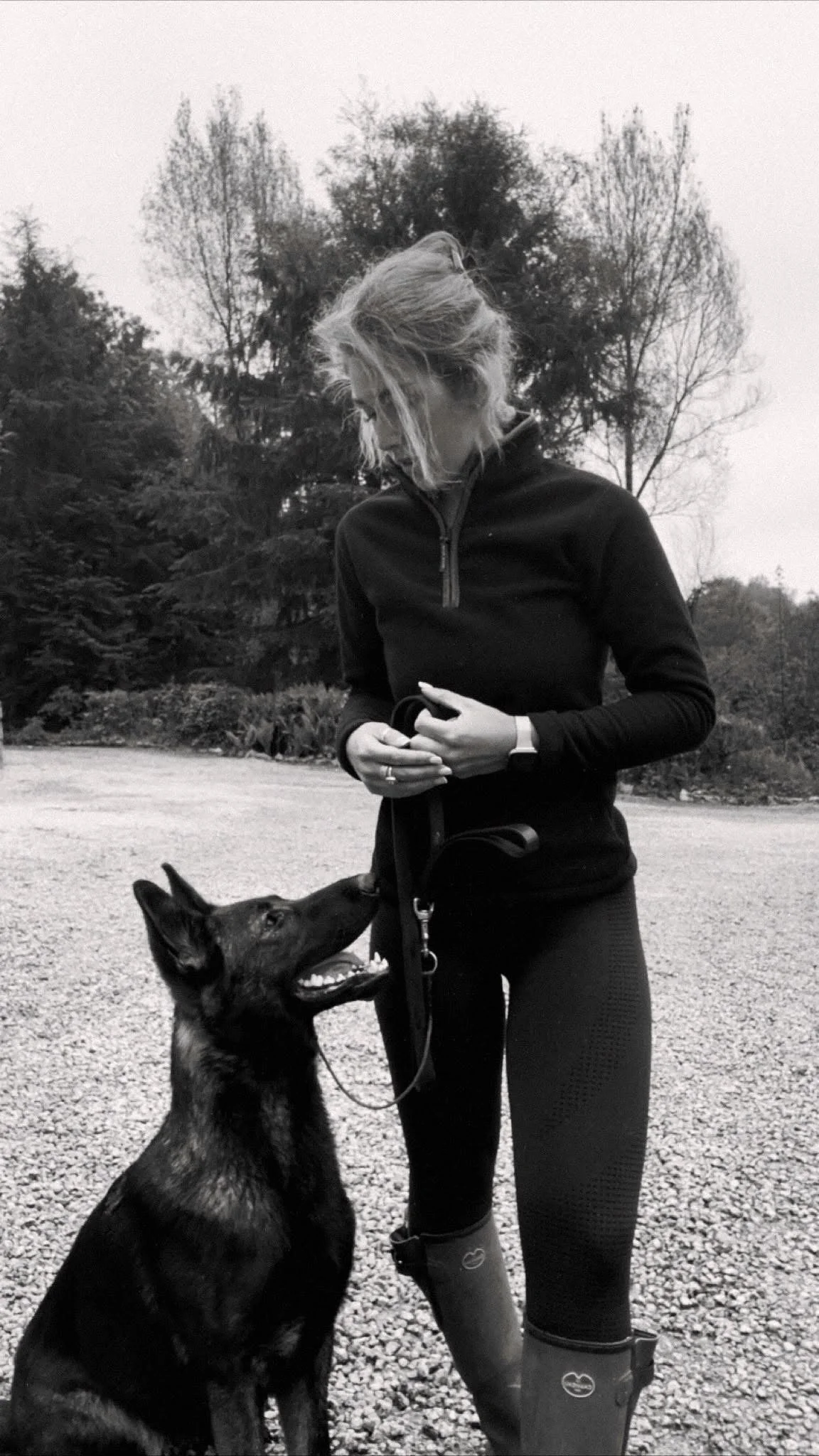 A woman outdoors wearing a black zip-up jacket and rubber boots, standing with a German shepherd dog on a leash, looking down at the dog in a natural setting with trees in the background.