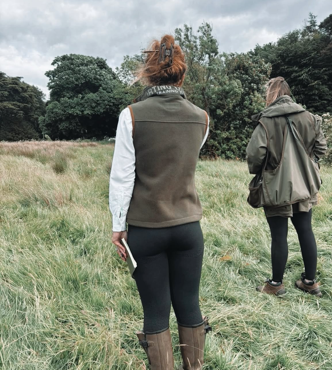 Two women walking through a grassy field on a cloudy day, wearing outdoor and hiking attire.