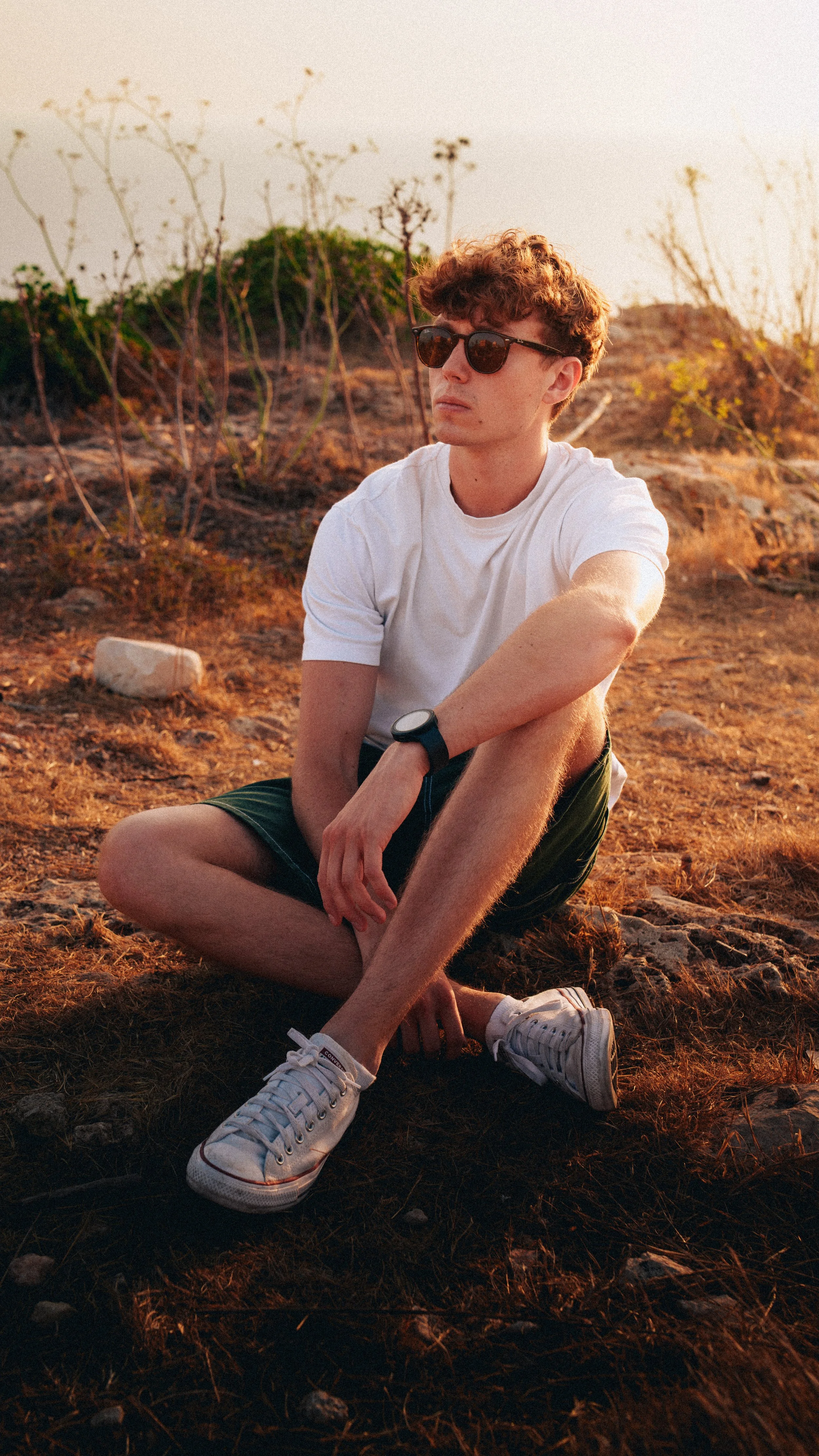 A young man with curly hair wearing sunglasses, a white t-shirt, dark shorts, white sneakers, and a watch sitting cross-legged on dry ground outdoors during sunset.