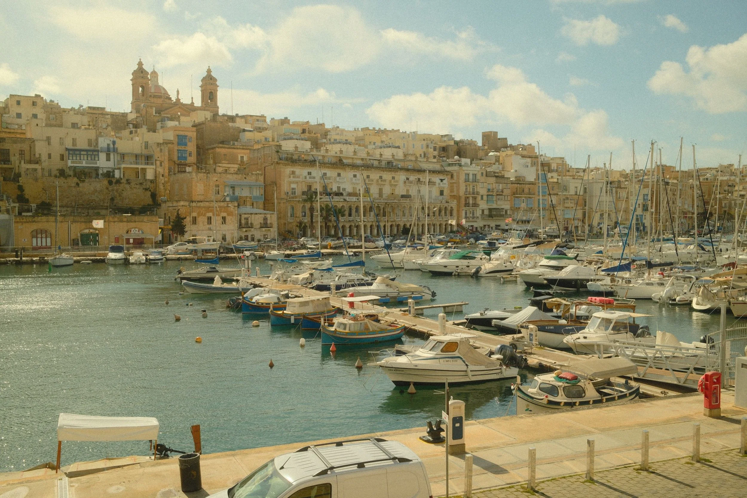 A harbor filled with boats and yachts docked in the water, with a hillside cityscape featuring historic buildings and a church with twin towers in the background, under a partly cloudy sky.
