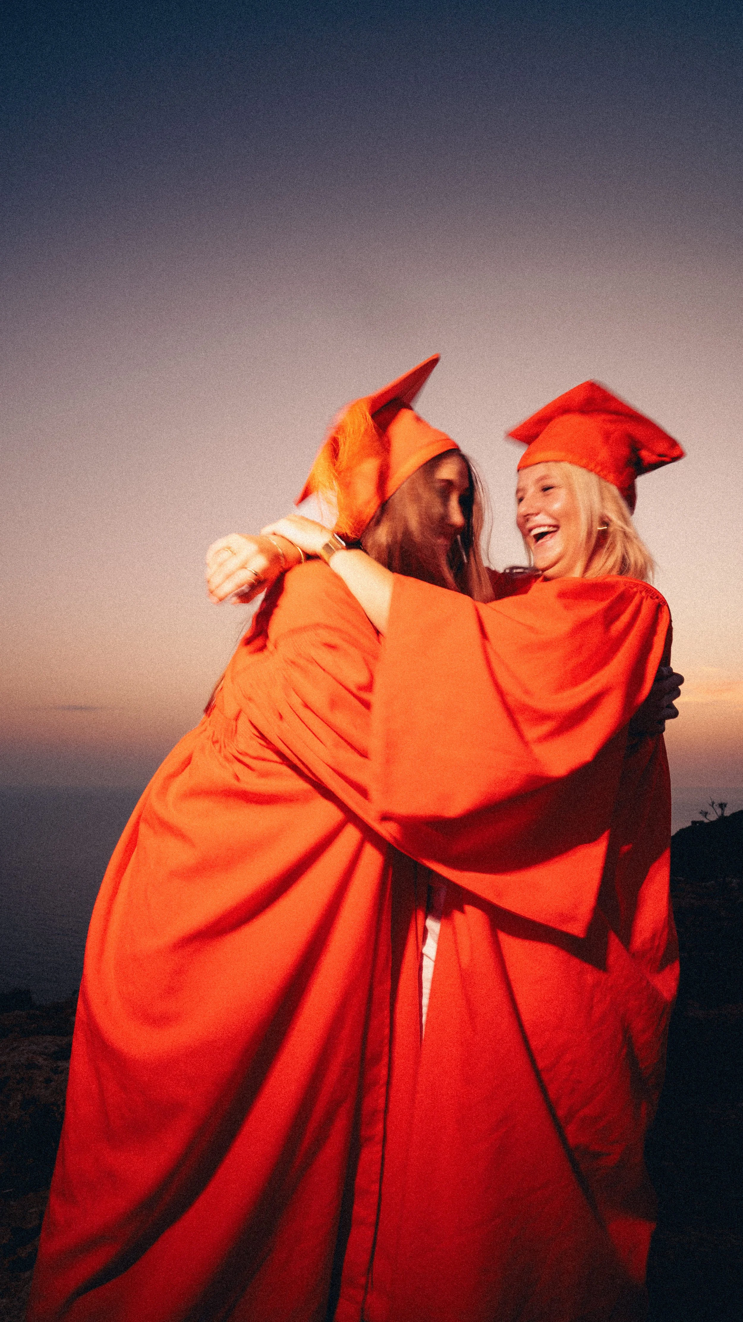 Two women wearing red graduation gowns and caps hugging outdoors during sunset.