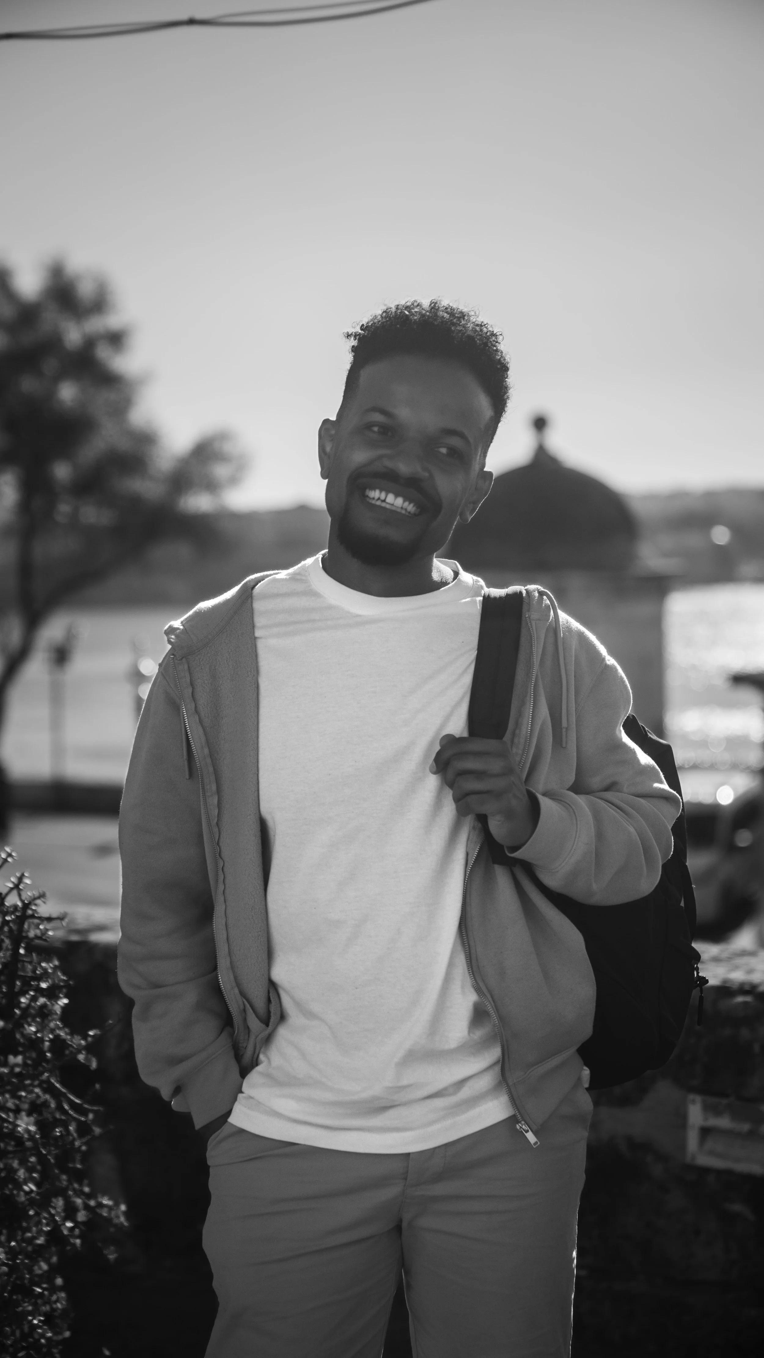 Black and white photograph of a smiling man with a beard, wearing a white t-shirt and a hoodie, carrying a backpack over one shoulder, standing outdoors near water with a blurred background including a tree and a dome-shaped structure.
