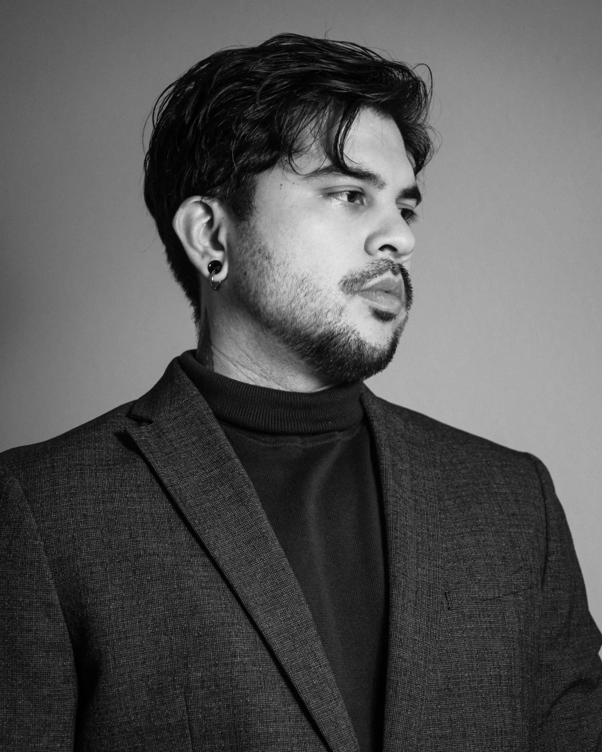 Schwarz-weiß Porträt eines Mannes mit dunklem Haar, Bart, Eiern, Ohrringen und dunklem Turtleneck, gekleidet in einem grauen Blazer, blickt nach rechts.