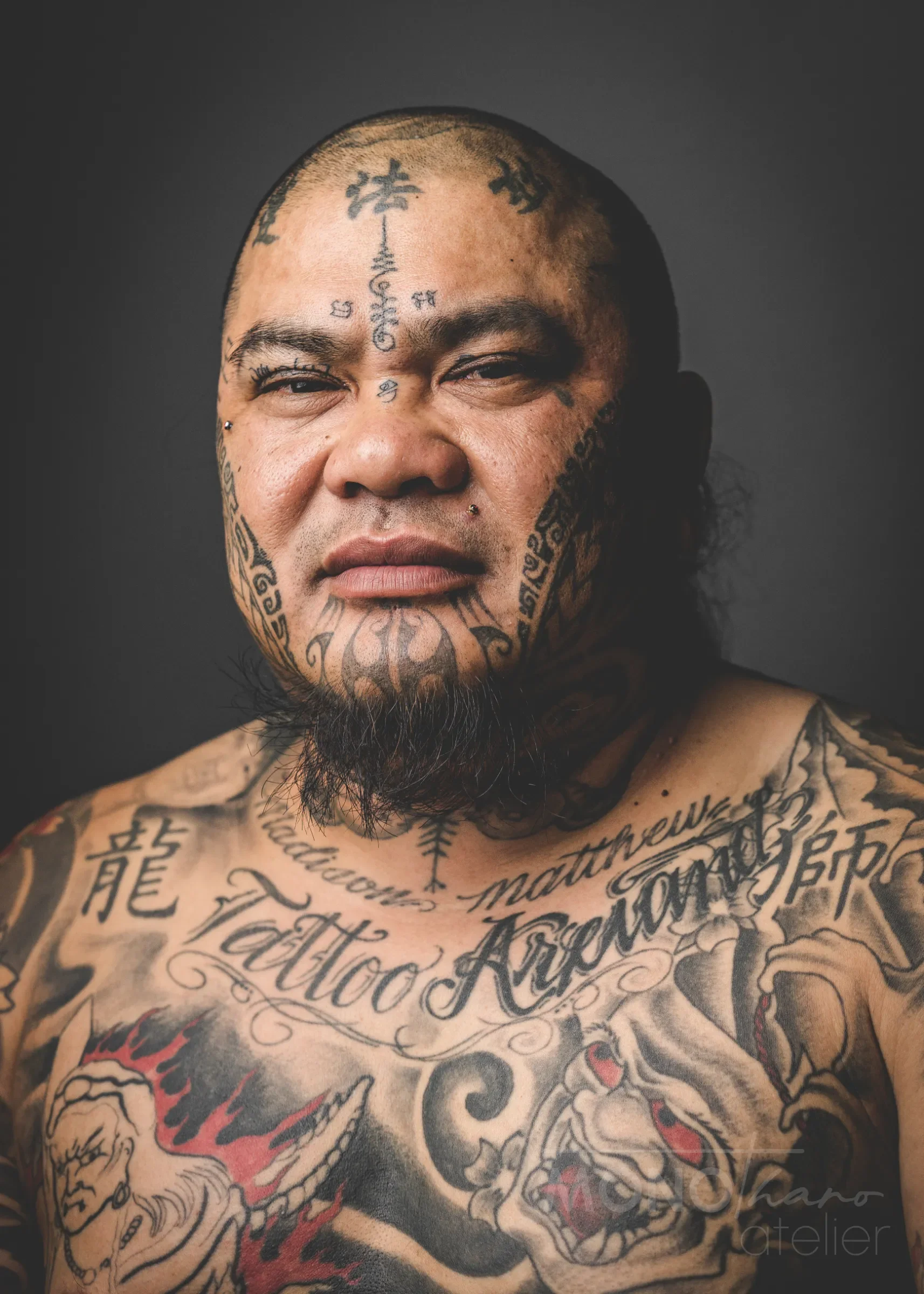 Nahaufnahme eines Mannes mit zahlreichen Tattoos im Gesicht, auf Brust und Gesicht. Er hat einen Bartschnitt und trägt keinen Schmuck. Der Hintergrund ist dunkel.